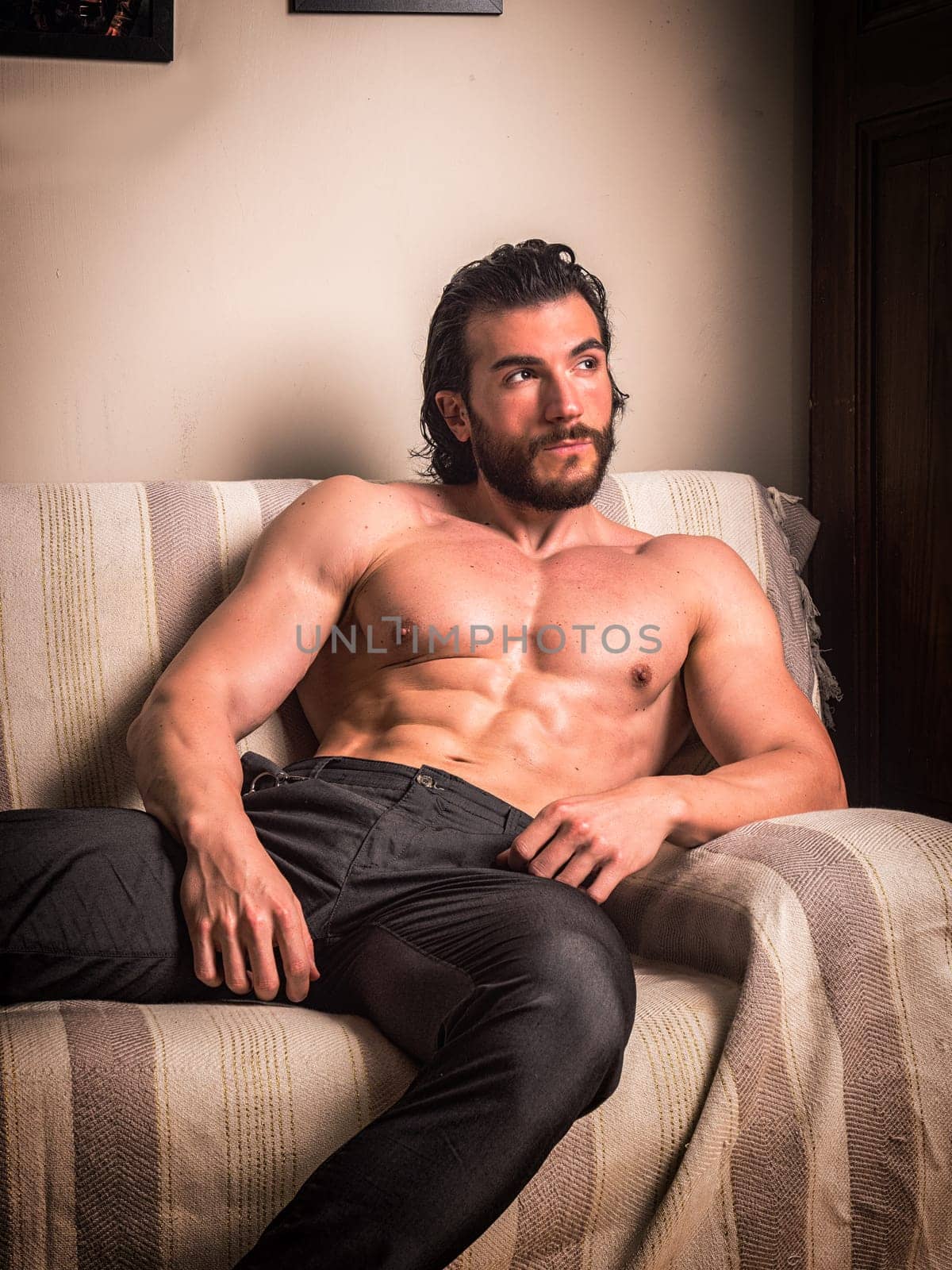 Handsome muscular young man at home laying on couch with shirt open on naked torso, an expression of tranquility, looking away, in a health and fitness concept