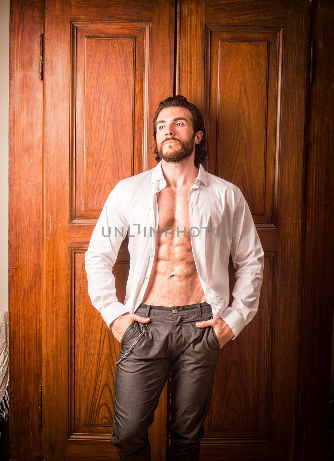 Sexy handsome man standing in white open shirt with a smile in front of wood closet doors