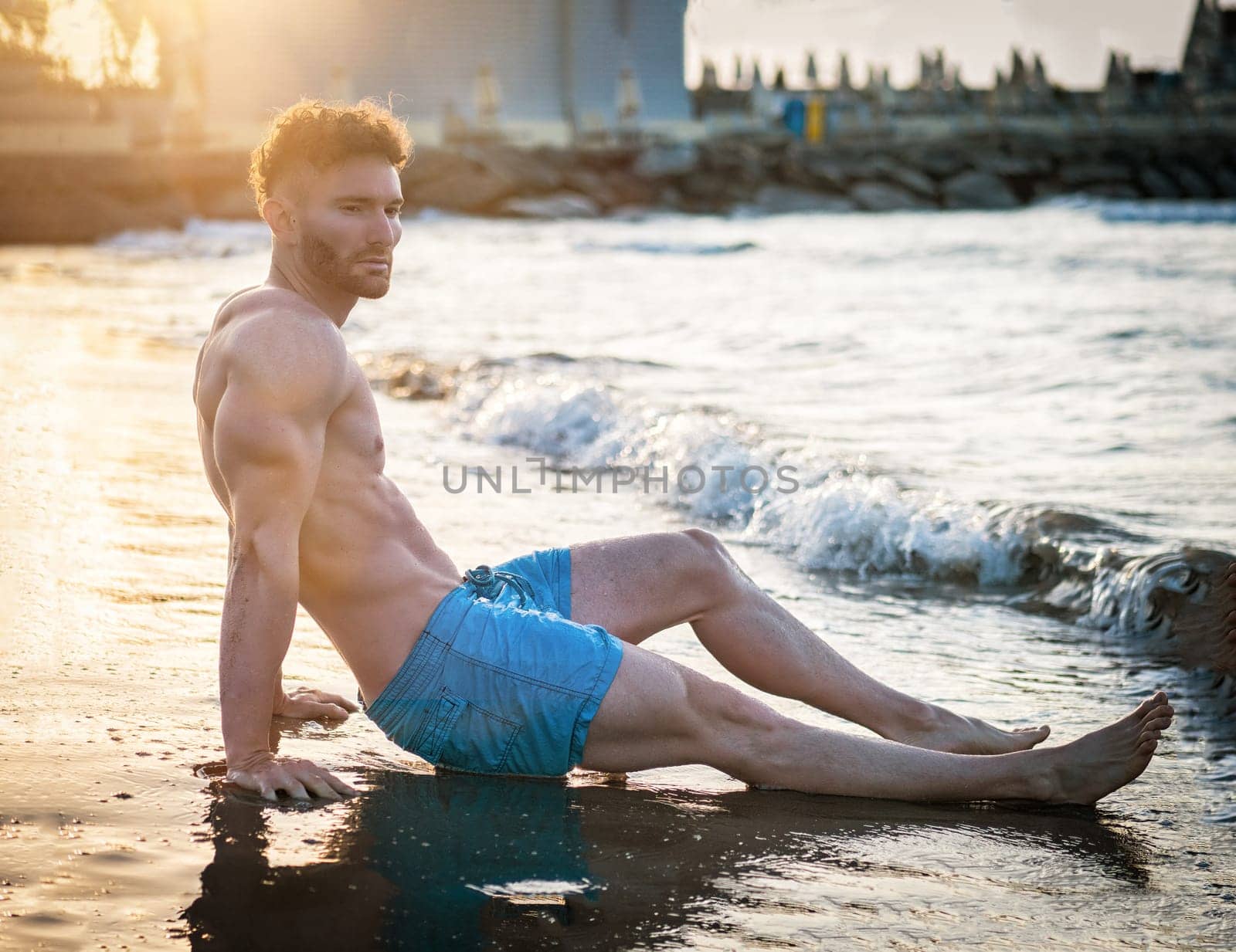 A shirtless muscular man sitting on a beach next to the ocean at dawn