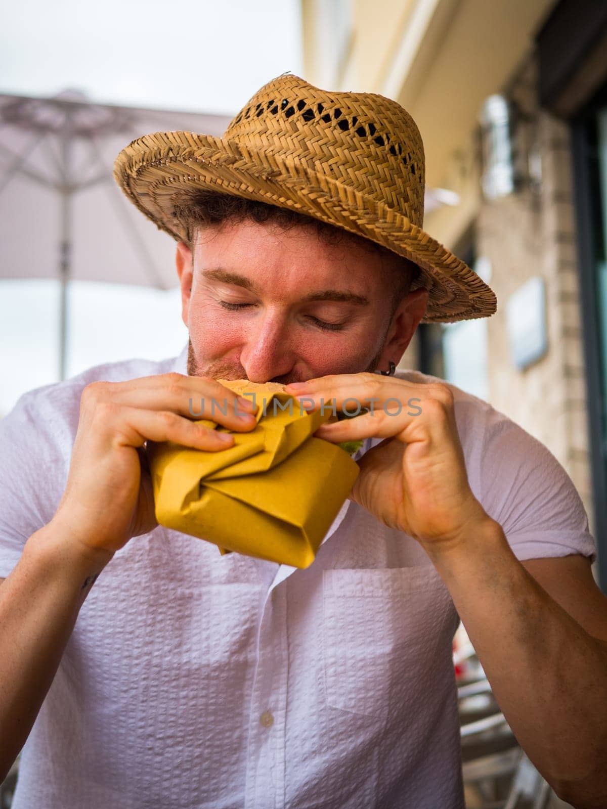 A man in a straw hat is holding a sandwich outdoor, sitting and looking at the food.