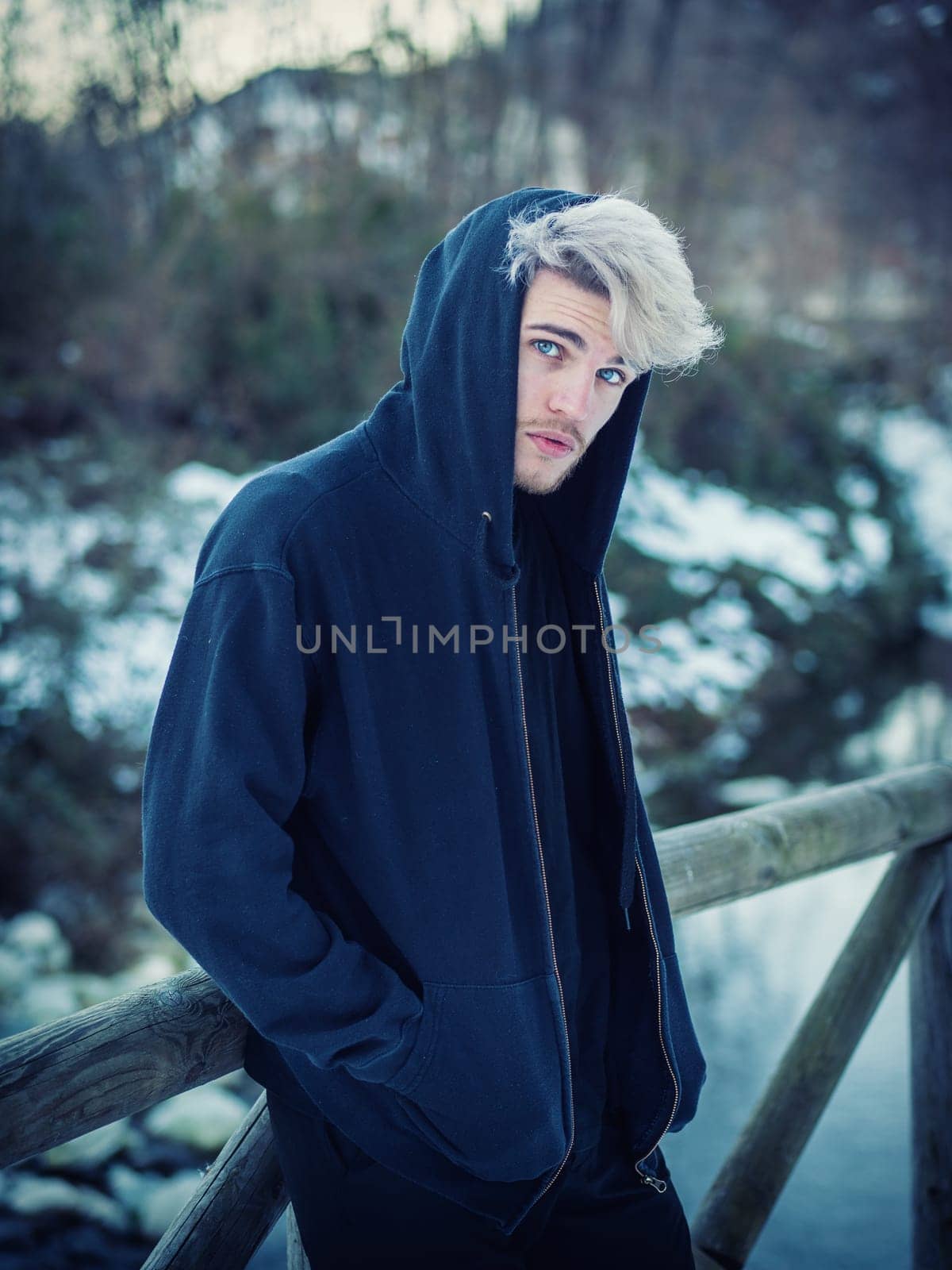Portrait of young man in hoodie posing outdoor in winter setting with snow all around, looking at camera