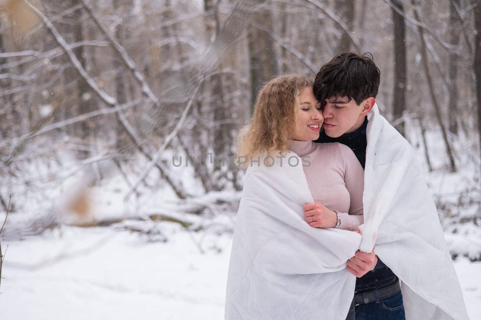 A young couple walks in the park in winter. The guy and the girl are kissing wrapped in a white blanket outdoors. by mrwed54
