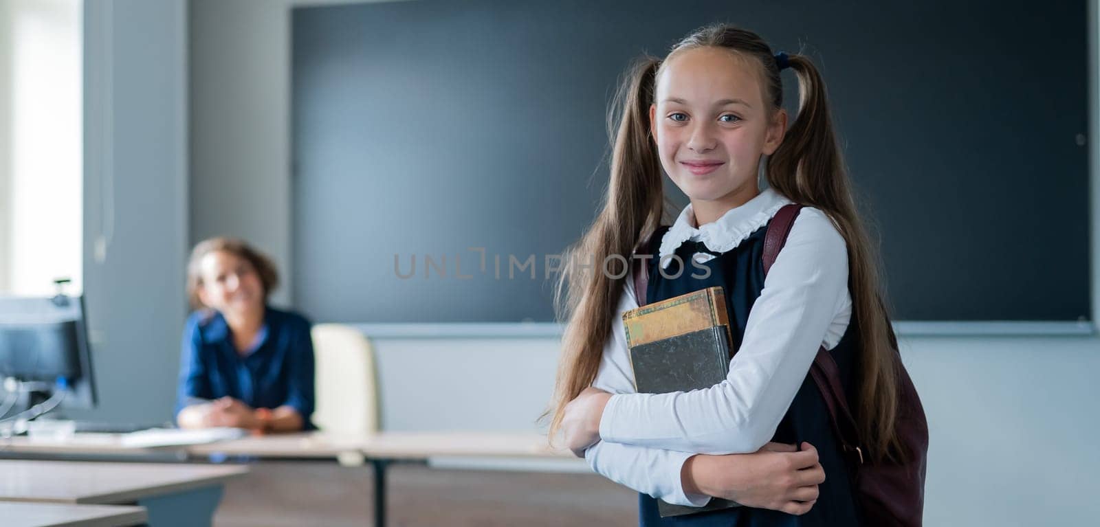 Caucasian girl and female teacher in the classroom. The schoolgirl is holding a textbook