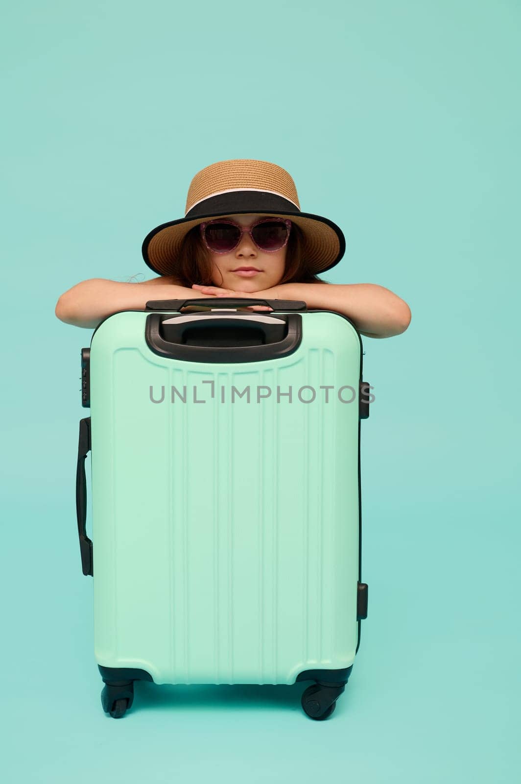 Cute child girl girl traveler in sunglasses and summer straw hat, going for weekend getaway, confidenty looking at camera, posing with blue suitcase, isolated over studio background. Copy ad space