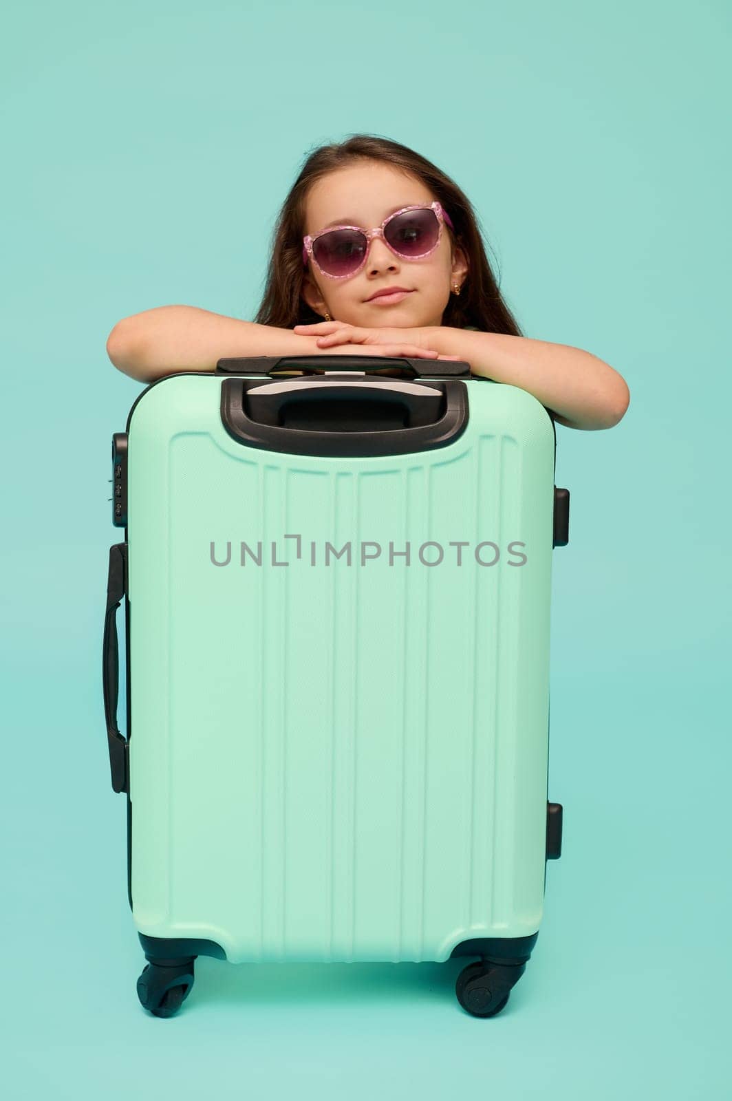 Adorable Caucasian little child girl traveler in trendy sunglasses, going for weekend getaway, confidenty looking at camera, posing with blue suitcase, isolated over studio background. Copy ad space