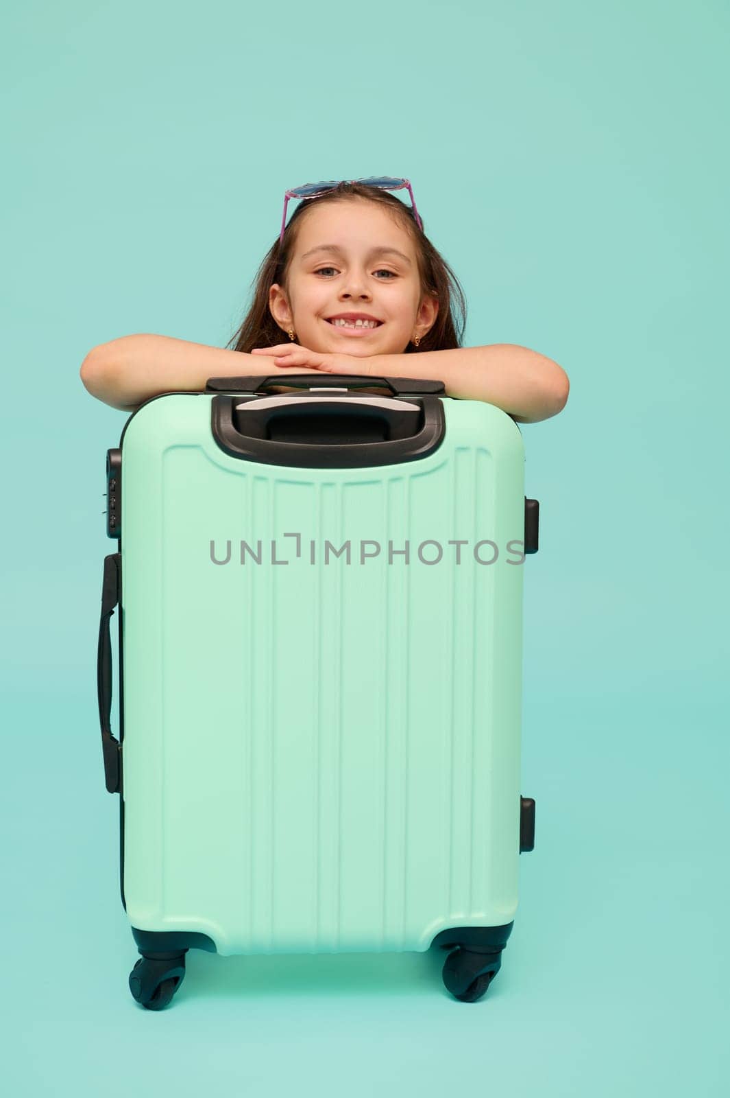 Adorable little child girl looking at camera, posing with blue suitcase, isolated over studio background. Copy ad space by artgf