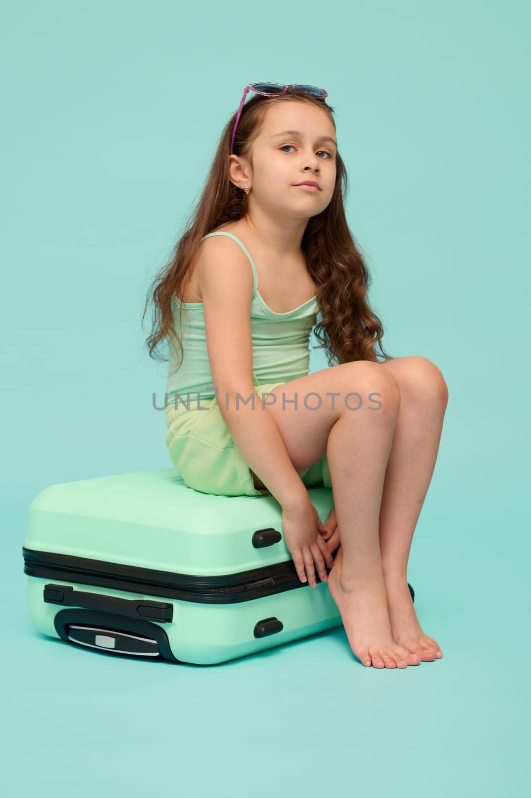 Full length vertical emotional portrait of a charming little kid girl in summer wear, sitting barefoot on her suitcase, smiling looking at camera, going for holidays, isolated blue studio background