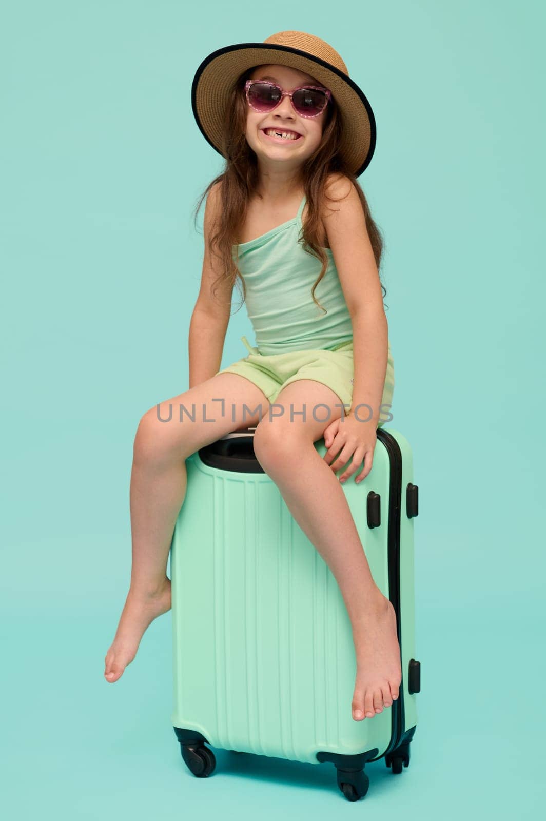 Full length vertical portrait of a Caucasian cute child girl 6 years old, sitting on her suitcase, isolated background by artgf