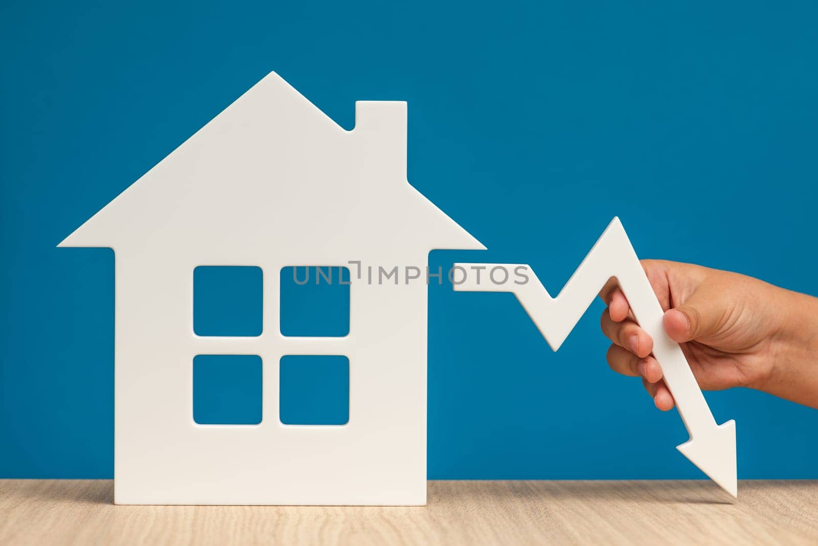 The collapse in real estate prices. Sale in the housing market. Reducing the value of real estate. Model of a house on a blue background and hand holding a white graph arrow pointing down