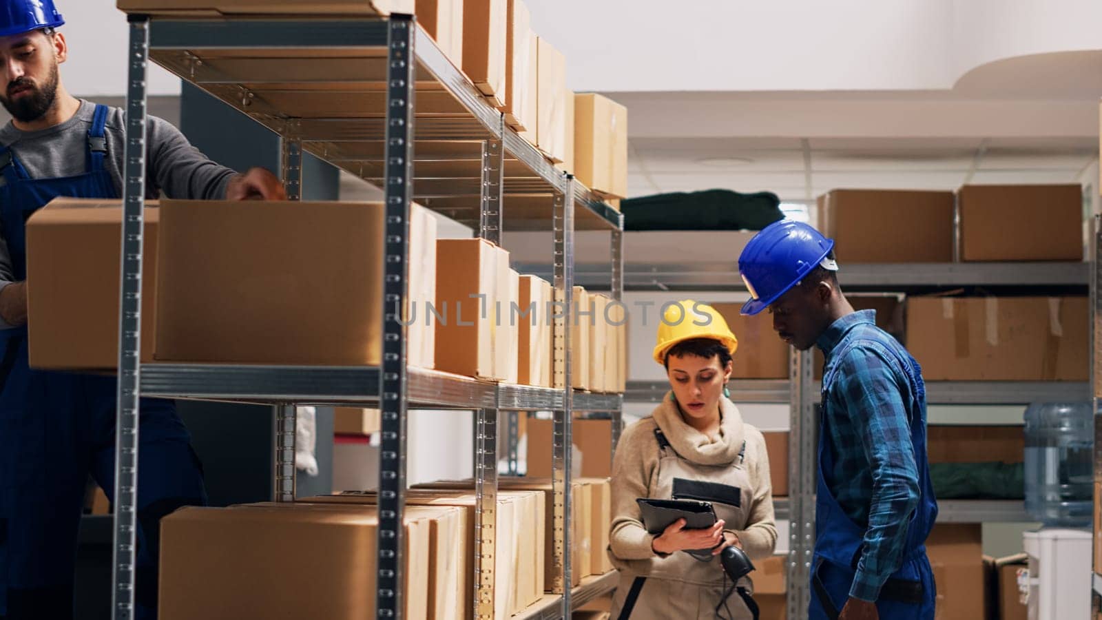 Diverse depot workers using digital tablet to scan goods, pointing scanner at barcodes on boxes to check stock logistics. Young employees in overalls working with cargo inventory.