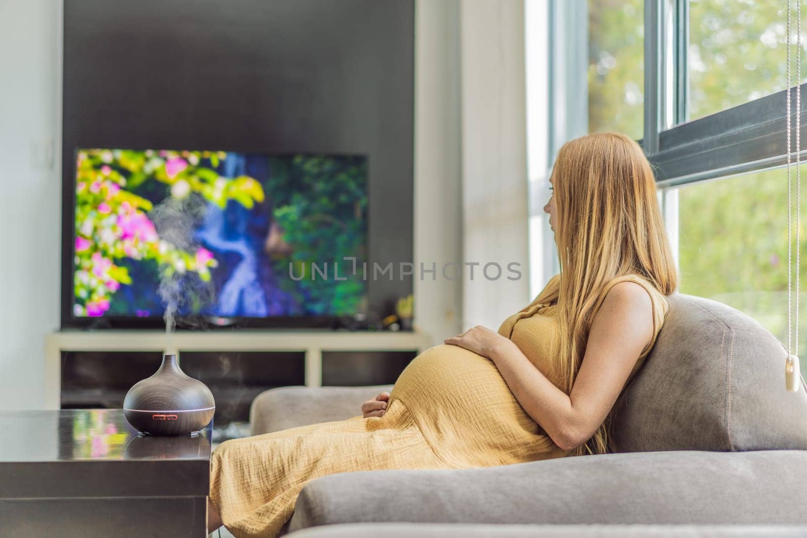 A blissful pregnant woman immerses in relaxation, savoring the soothing aroma from a diffuser while indulging in a calming TV video, embracing tranquility during her pregnant journey.