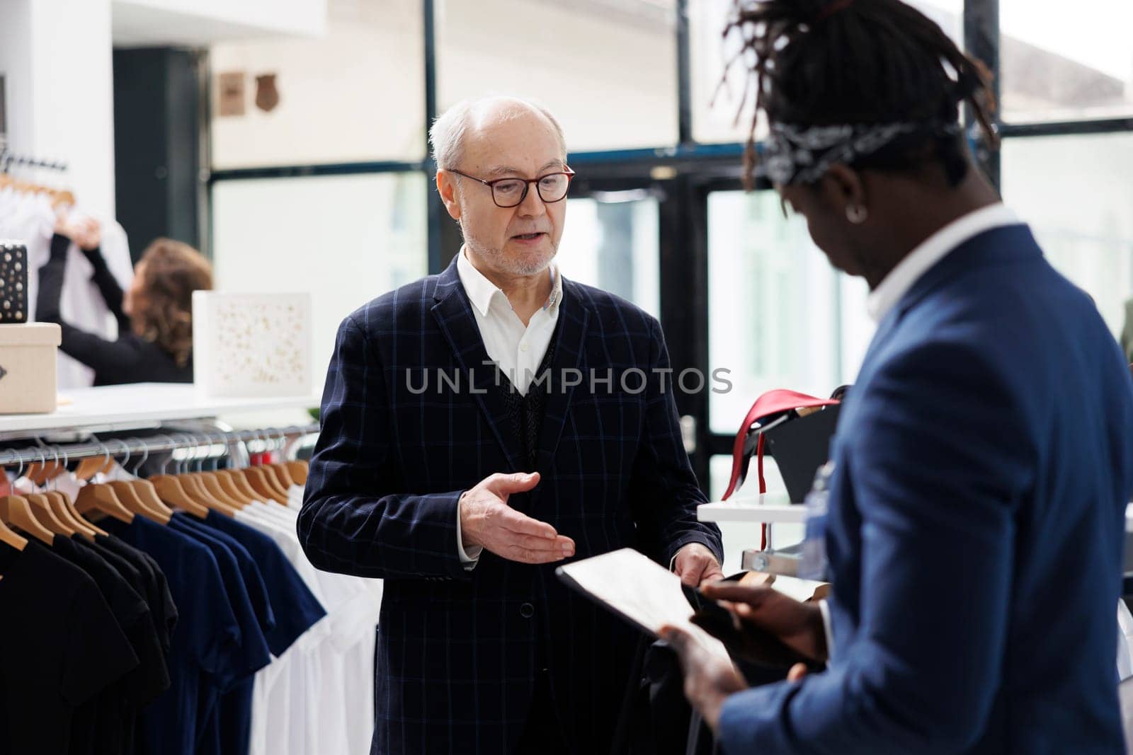 Elderly man asking worker for help with formal wear, african american employee checking merchandise inventory on tablet computer in showrrom. Senior customer looking at new fashion collection