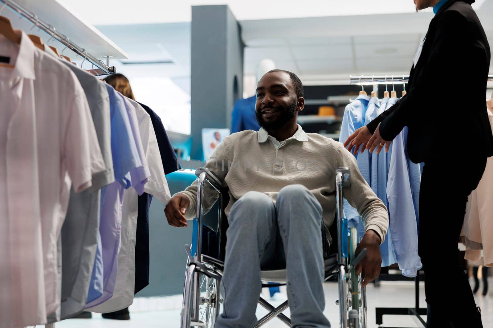 Smiling african american man in wheelchair getting assistance from clothing store employee while shopping for apparel. Menswear boutique customer with disability choosing outfit