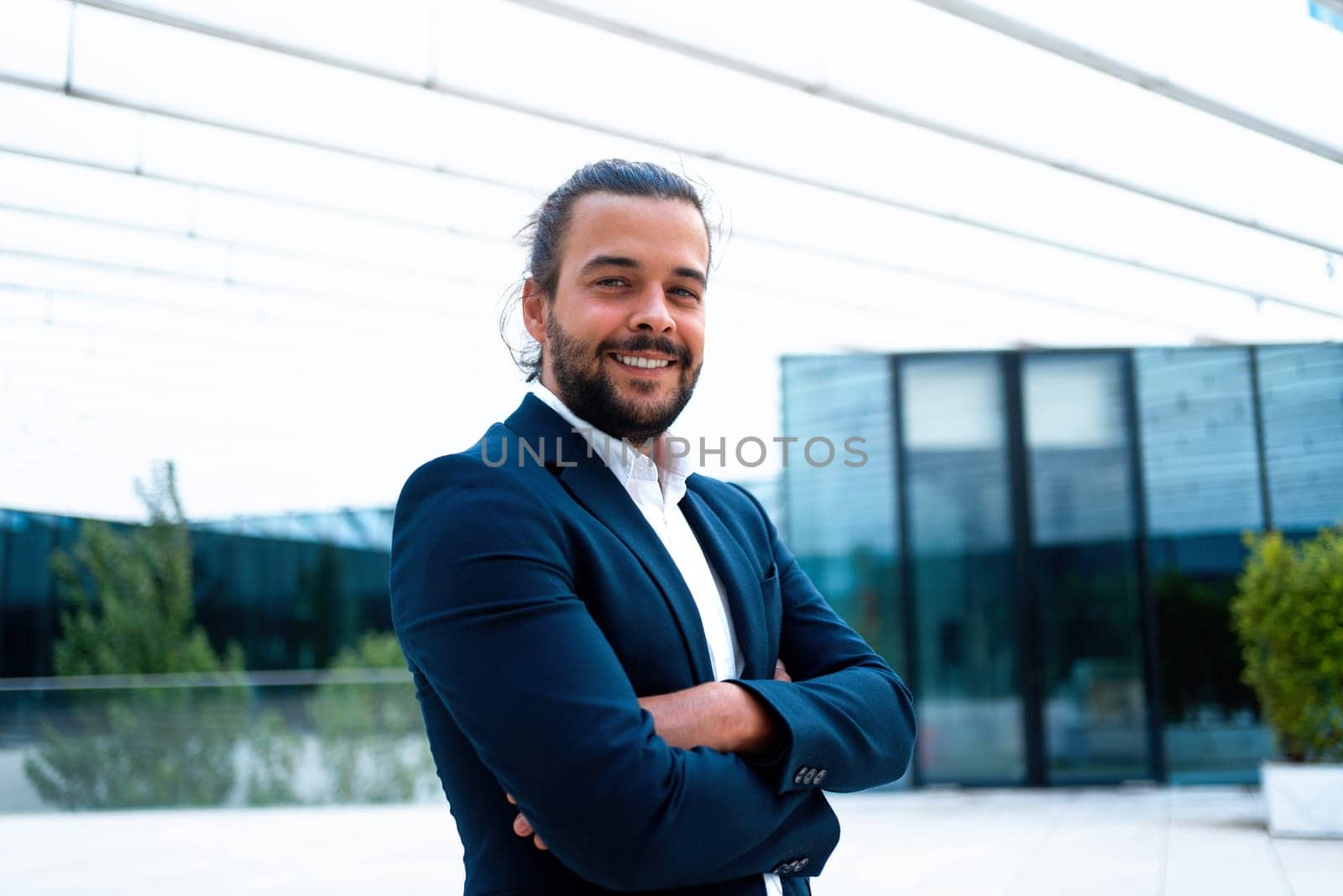 Successful businessman suit with beard standing in front of office glass building arm crossed confidently looking in camera and smile. Hispanic male business person portrait. Free space