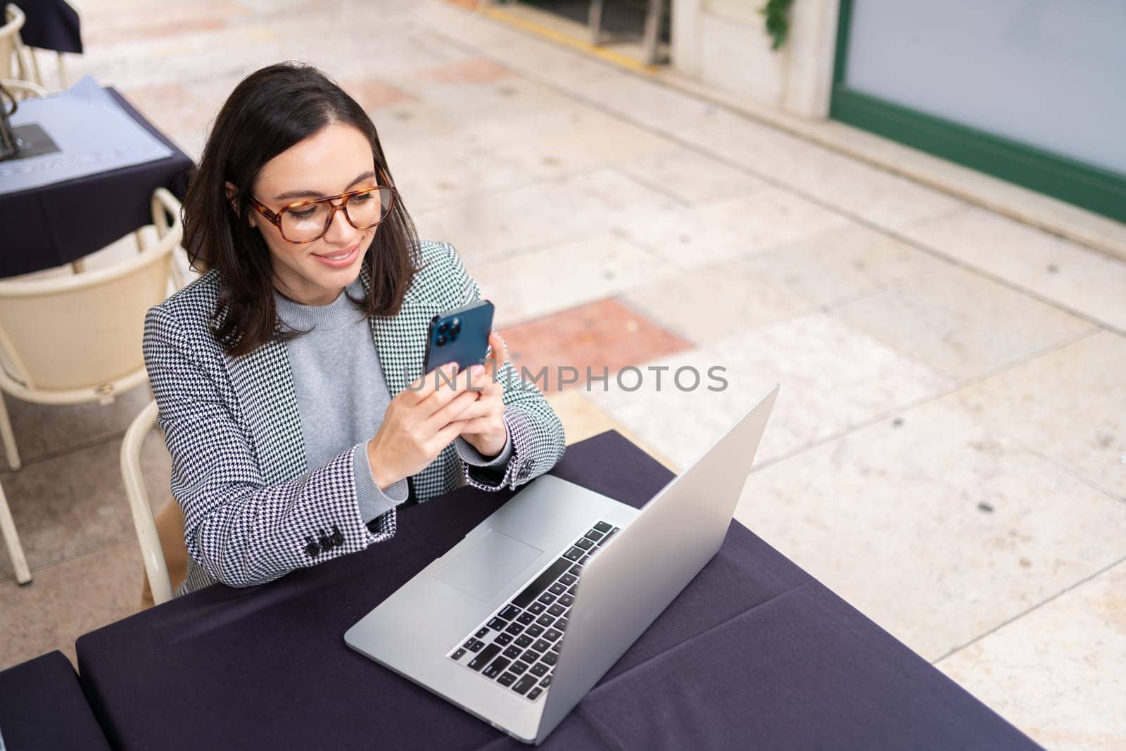 Brunette woman in casual clothes working with laptop using mobile phone at cafe. Freelancer using smartphone and laptop sitting outdoor cafe. High angle view, copy space