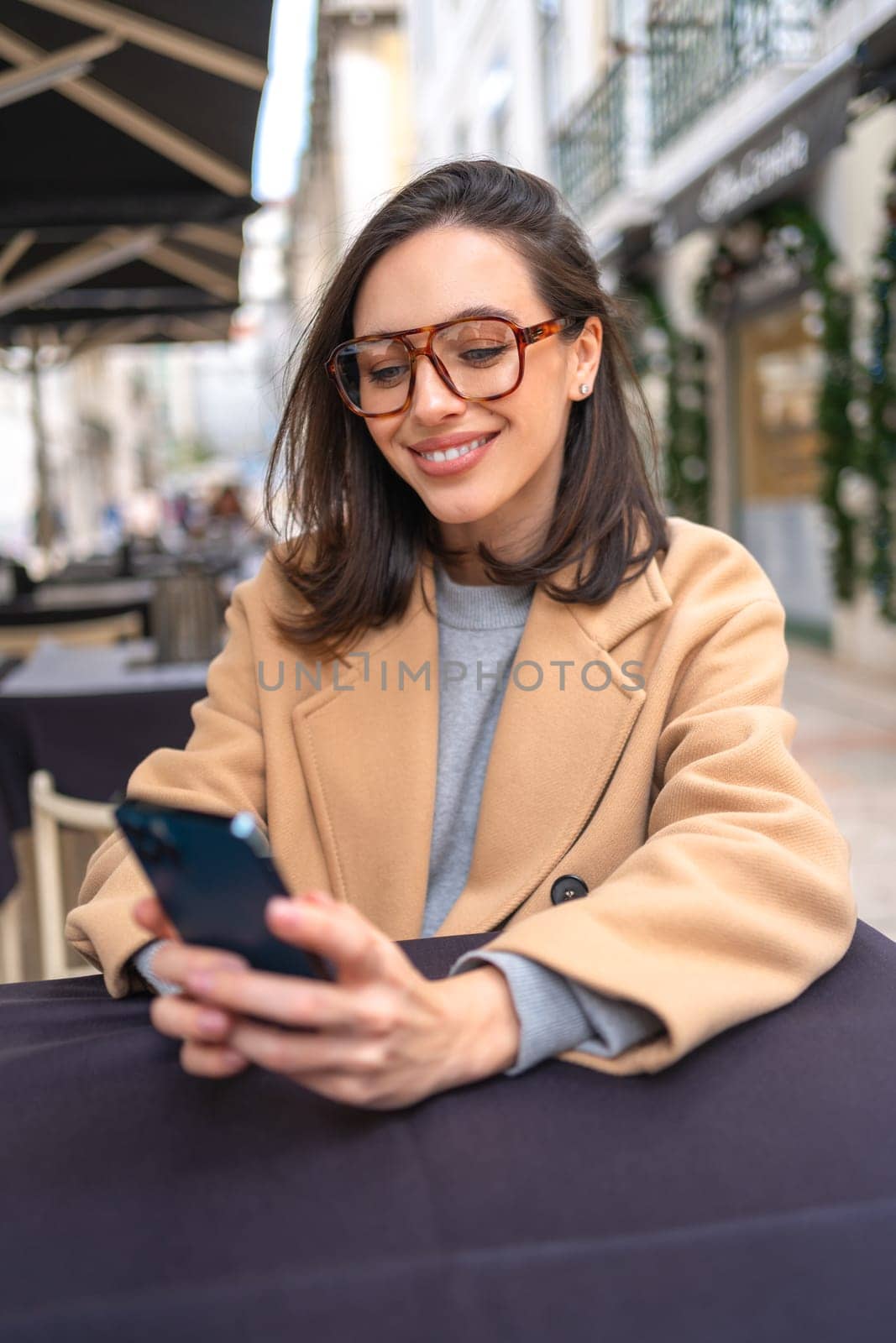 Happy middle-aged woman using smartphone sitting outdoor cafe. Smile businesswoman in glasses dressed trench coat holding smartphone in hand looking screen