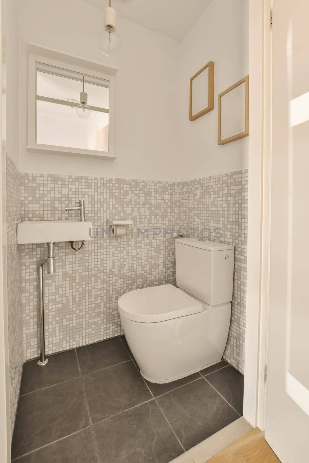 a white toilet in a small bathroom with grey tiles on the walls and floor to the right is a mirror