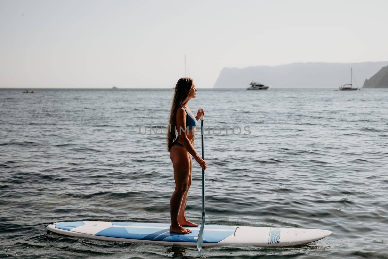 Woman sea sup. Close up portrait of happy young caucasian woman with long hair looking at camera and smiling. Cute woman portrait in a blue bikini posing on sup board in the sea by panophotograph