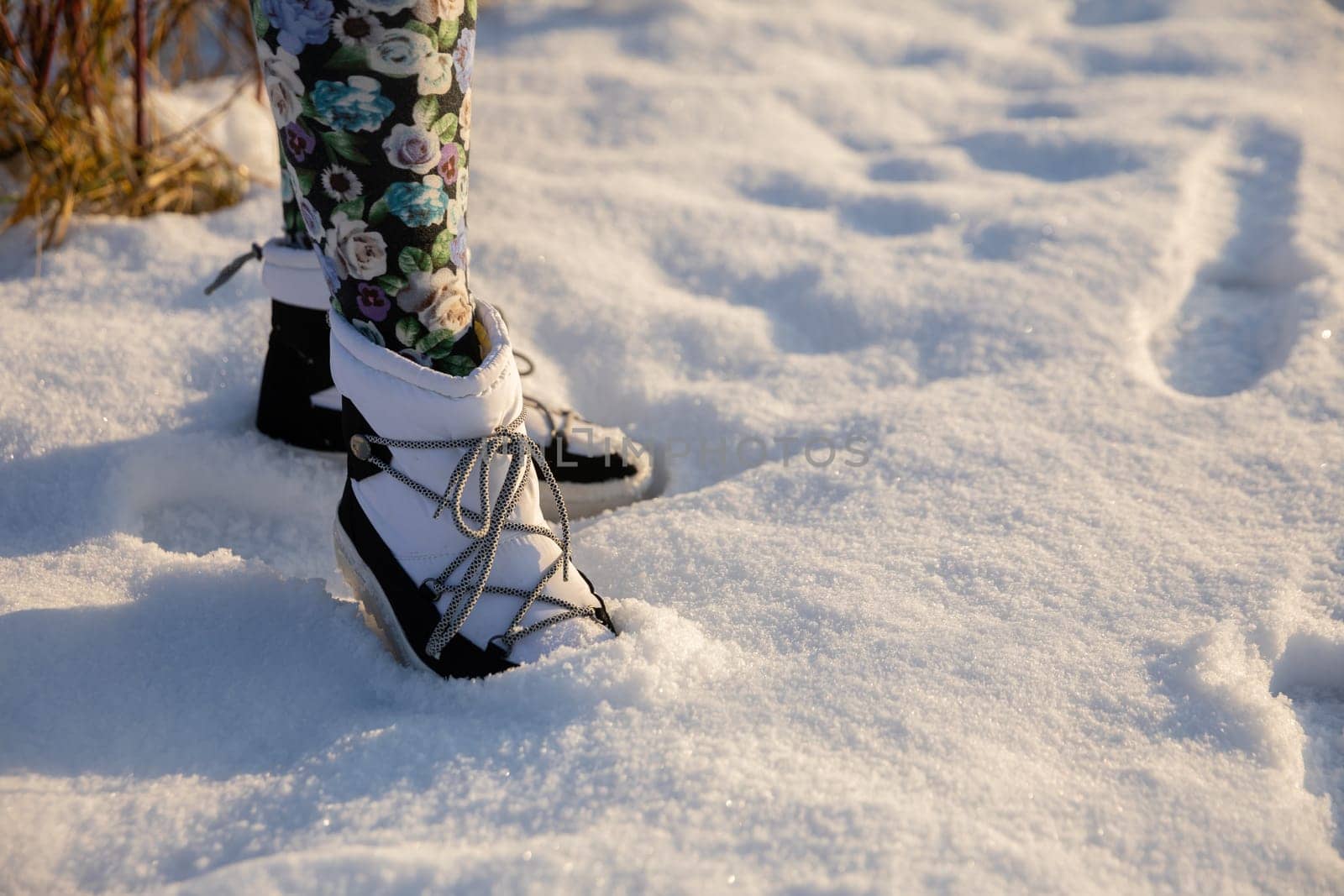 The person standing in the snow has one leg bent. She has the tip of her shoe buried in the snow. The woman is wearing white insulated boots with an upper and patterned leggings. A view of her legs from the knees down.