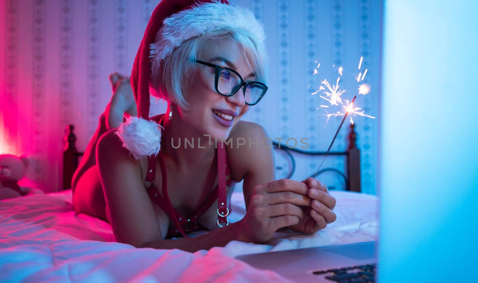 Sexy girl in lingerie wearing a Santa Claus hat poses sexily on a bed with a laptop by a Bengal fire on Christmas Day in neon lights