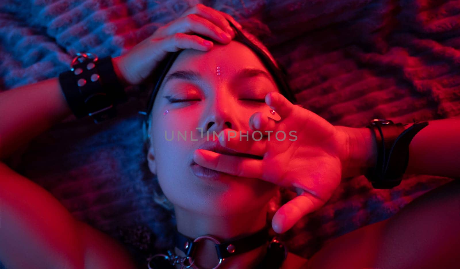 portrait of a sexy girl in a leather mask and bdsm accessories on a bed enjoying sex in orgasm with emotions on her face in neon light