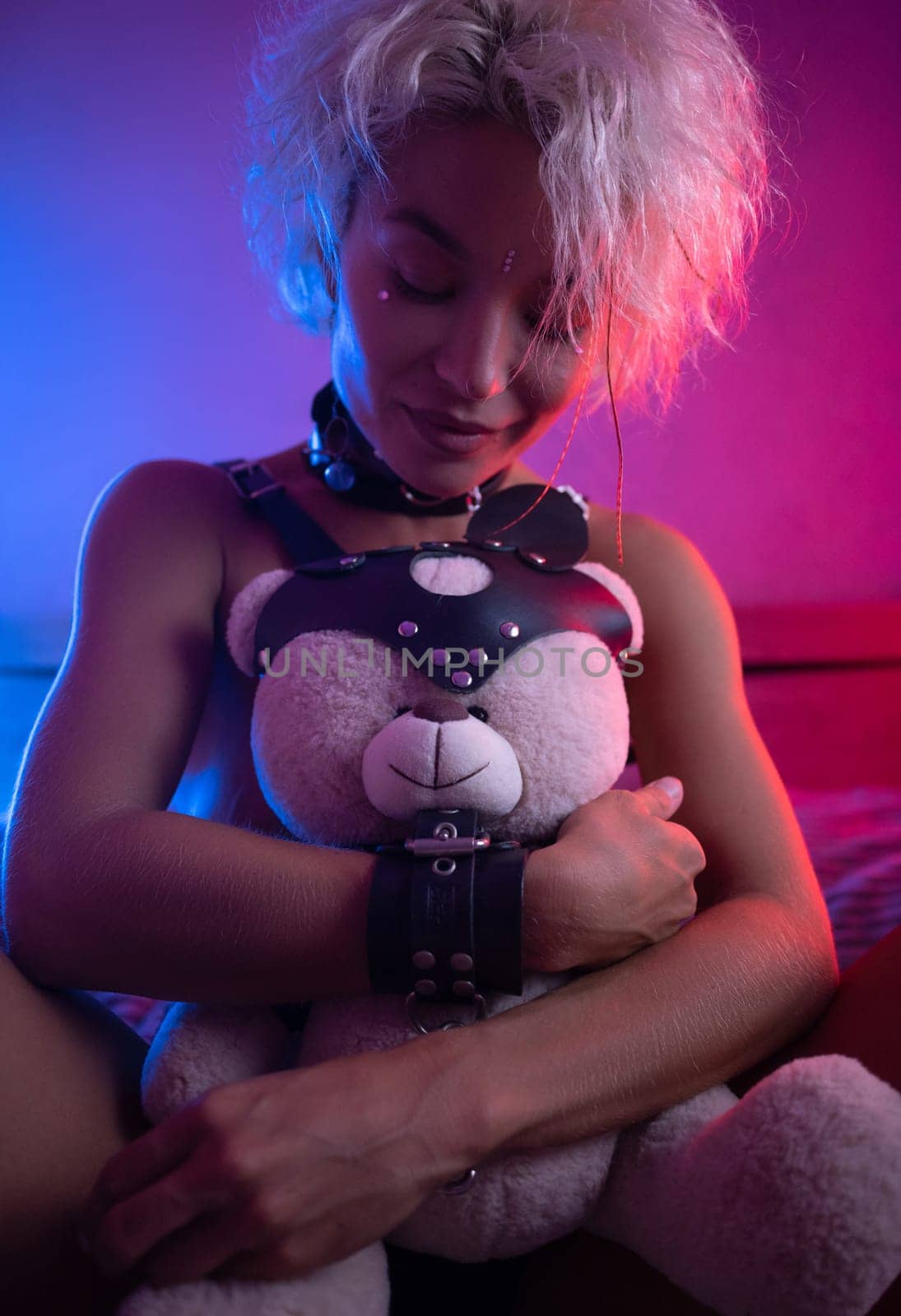 sexy girl in leather in a bdsm accessory on the bed with a cute teddy bear with emotions and in neon light