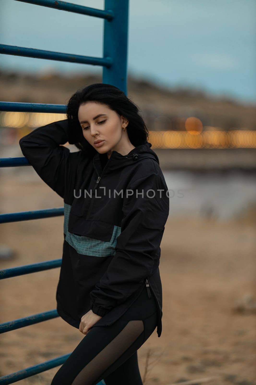 A beautiful brunette fitness model is working out on a sports ground by the sea. Young woman posing in an anorak and leggings outdoors. Fashionable sportswear for street training