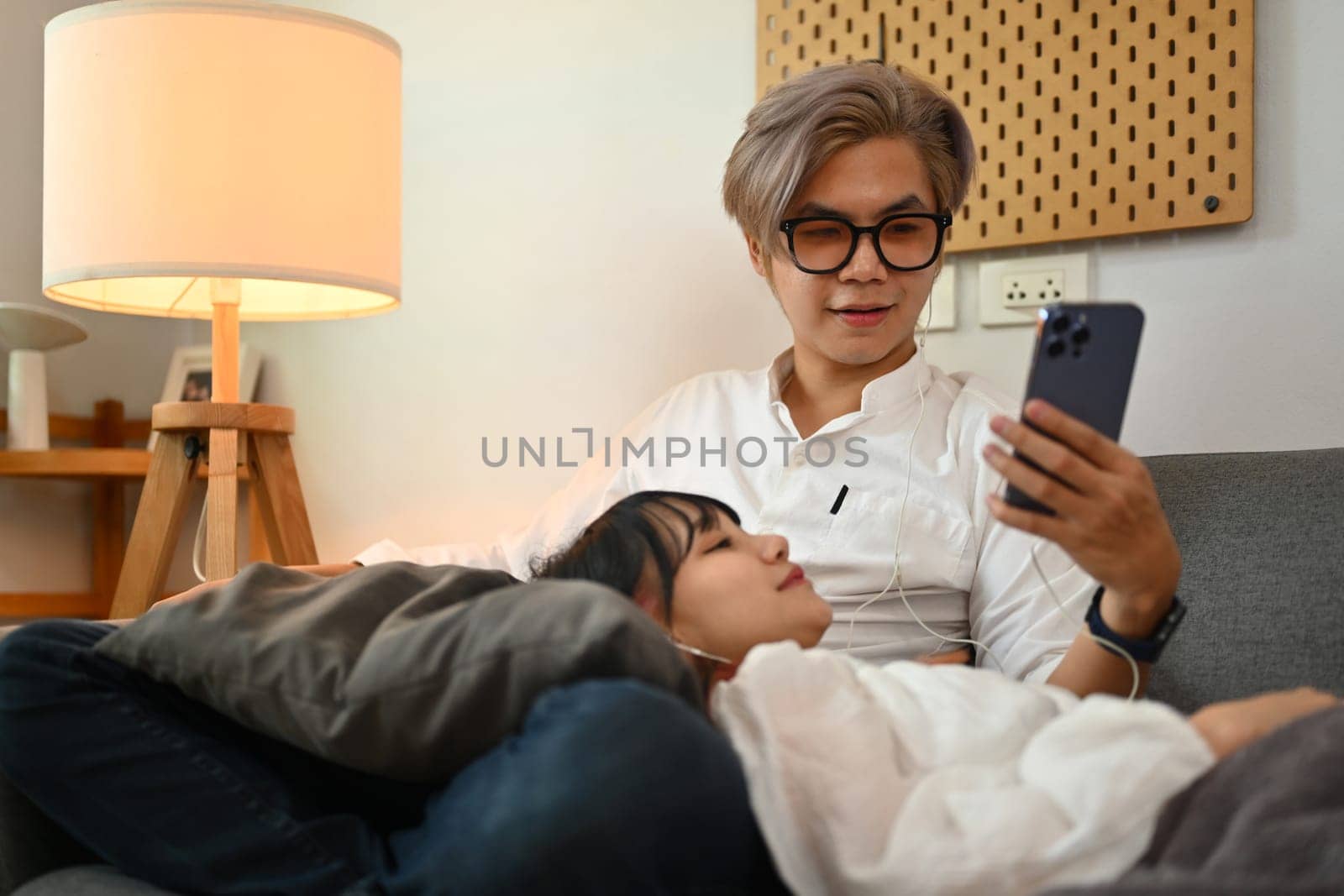 Smiling young asian man using mobile phone while relaxing with girlfriend on sofa at home.