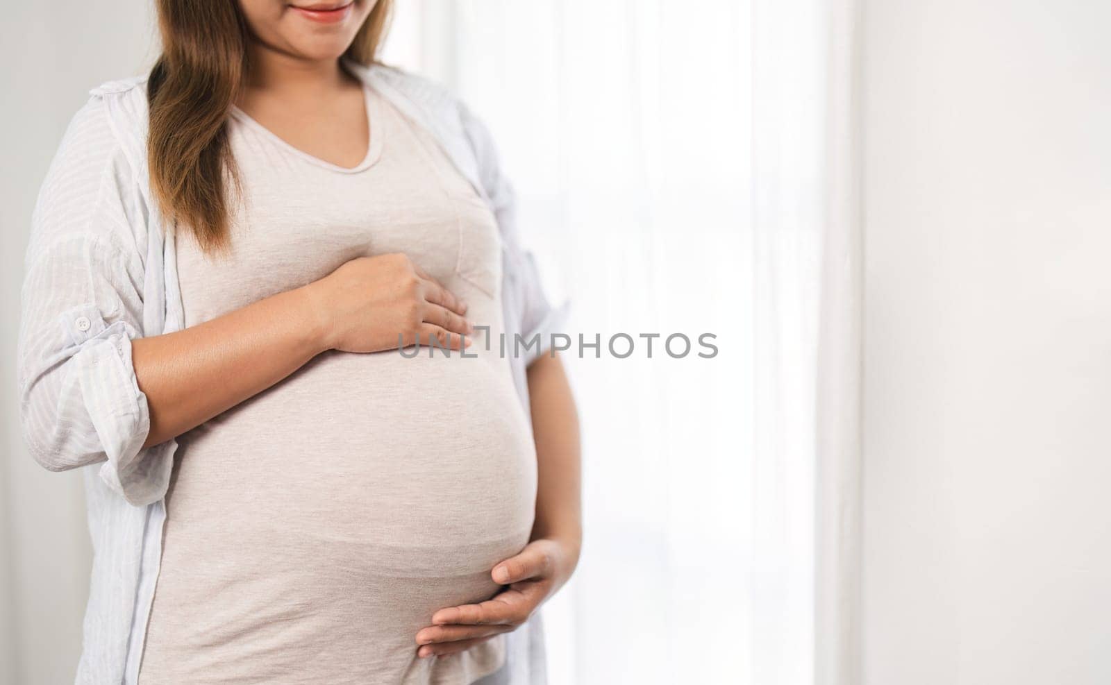 close up of a beautiful young woman during the third trimester of pregnancy Pregnant woman with arms on round belly, smiling, relax expression. by wichayada