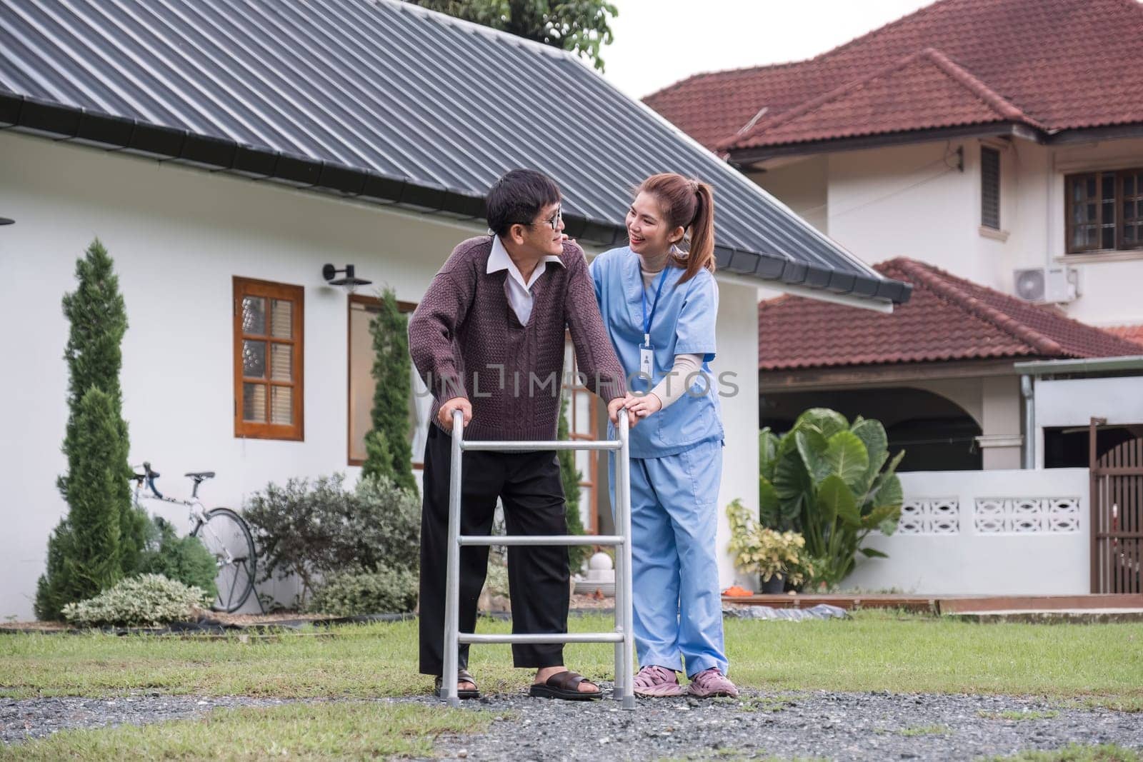 Asian female caregiver helps senior man walk A senior using a walker is assisted by a nurse at home. A nurse helps an elderly patient at a nursing home. Elderly care and healthy lifestyle.