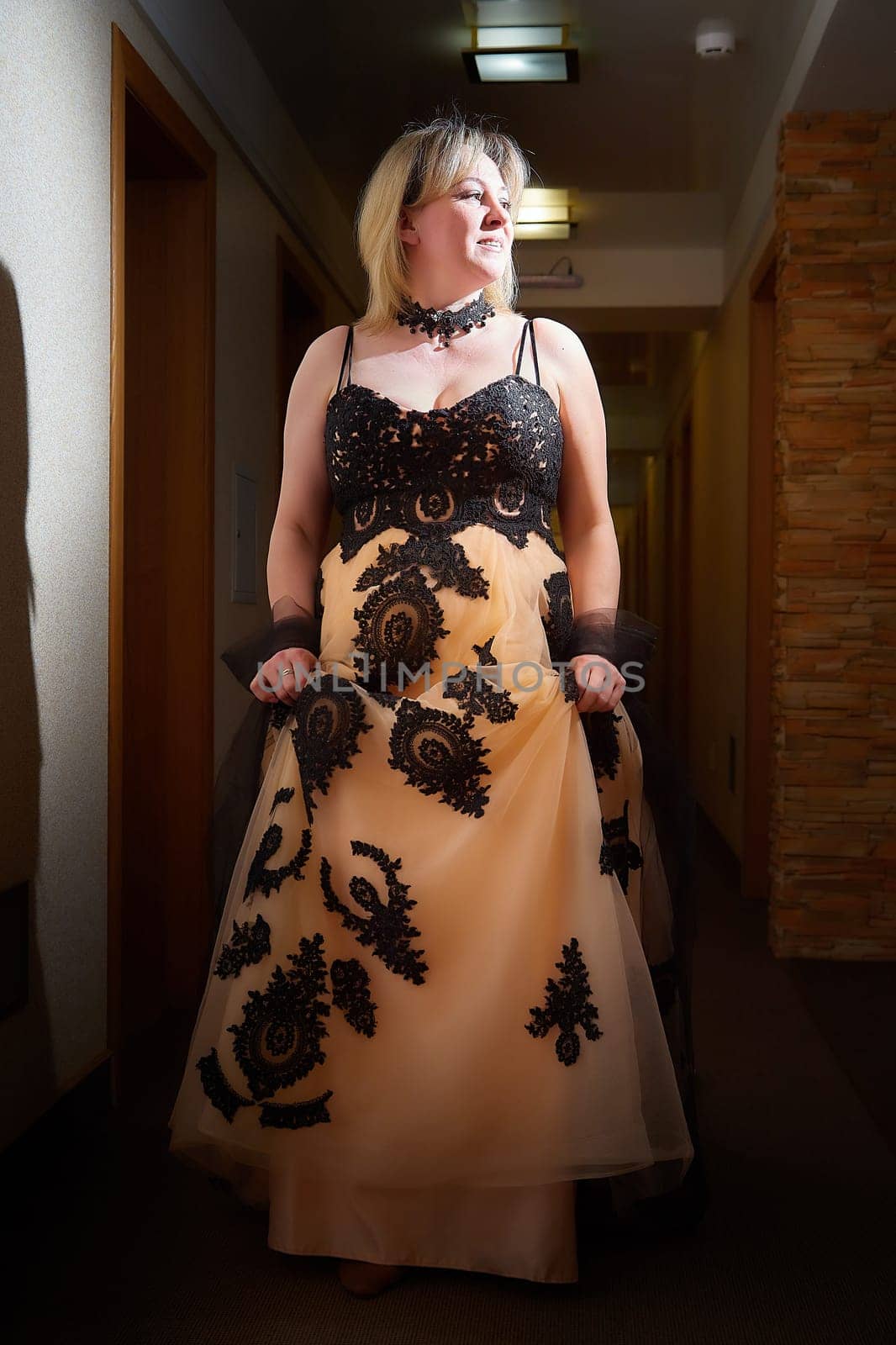 Full length view of a blonde female model, dressed in a black and white queen dress in dark corridor. Mysterious stranger alone inside castle or hotel