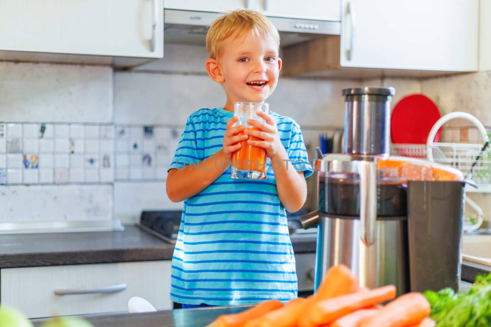 Happy smiling child enjoying freshly squeezed carrot juice in a cozy kitchen at home. Health and good mood with proper nutrition