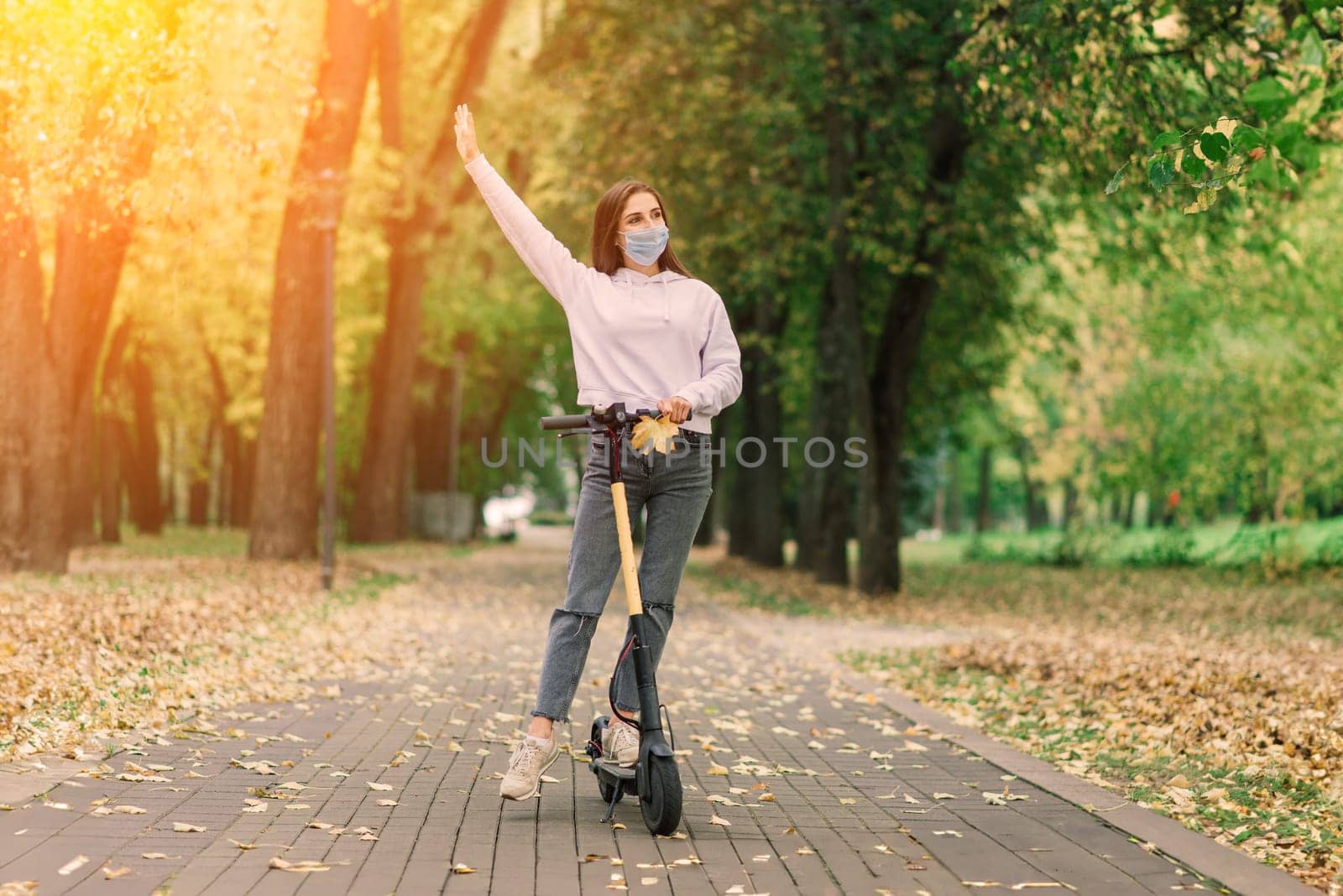 Casual caucasian female wearing protective face mask riding urban electric scooter in city park during covid pandemic. Urban mobility concept.