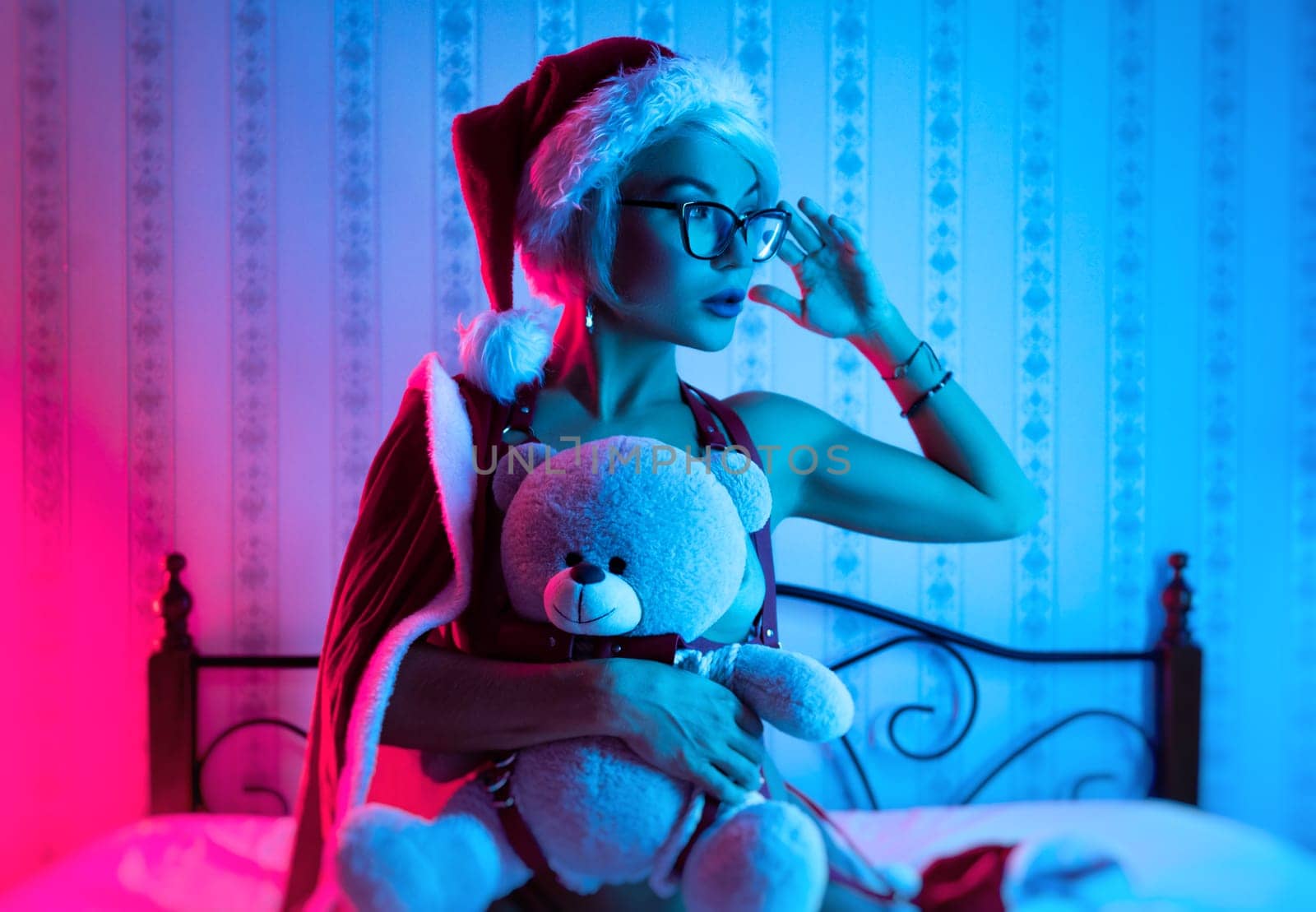 Sexy girl in underwear and a Santa Claus hat poses sexy on a bed with a teddy bear on a Christmas holiday in neon light