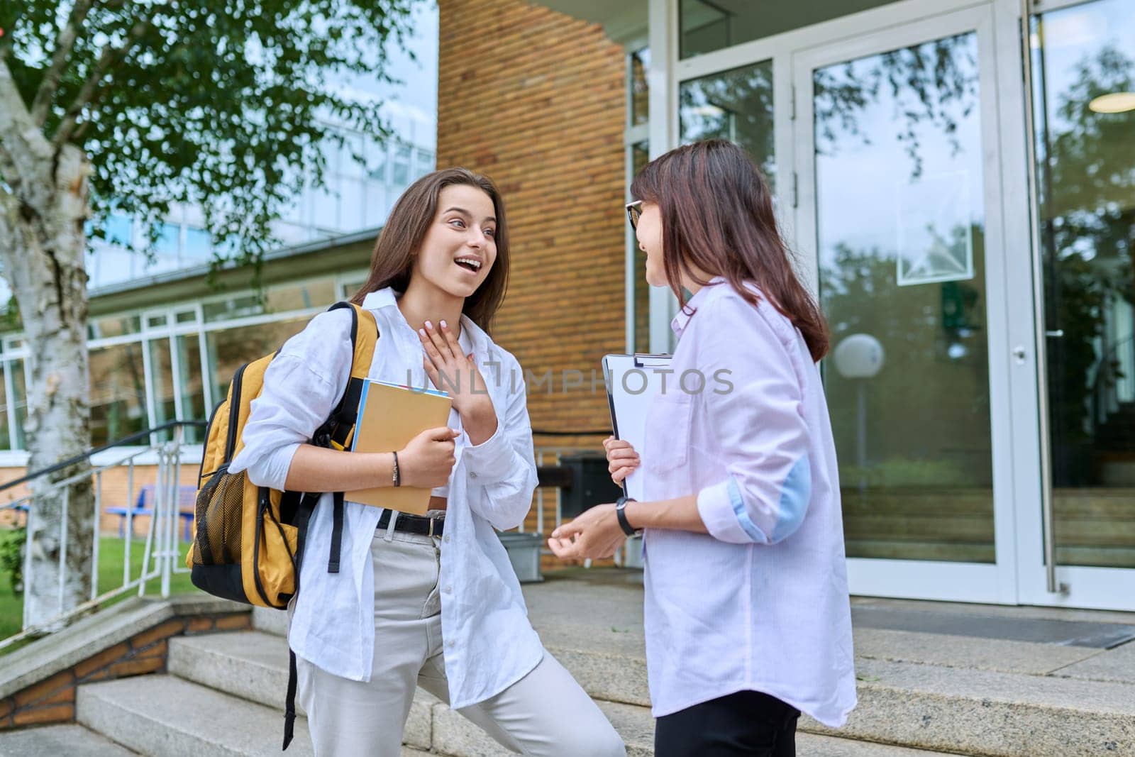 Teenage girl student talking to female teacher, standing on steps of educational building by VH-studio