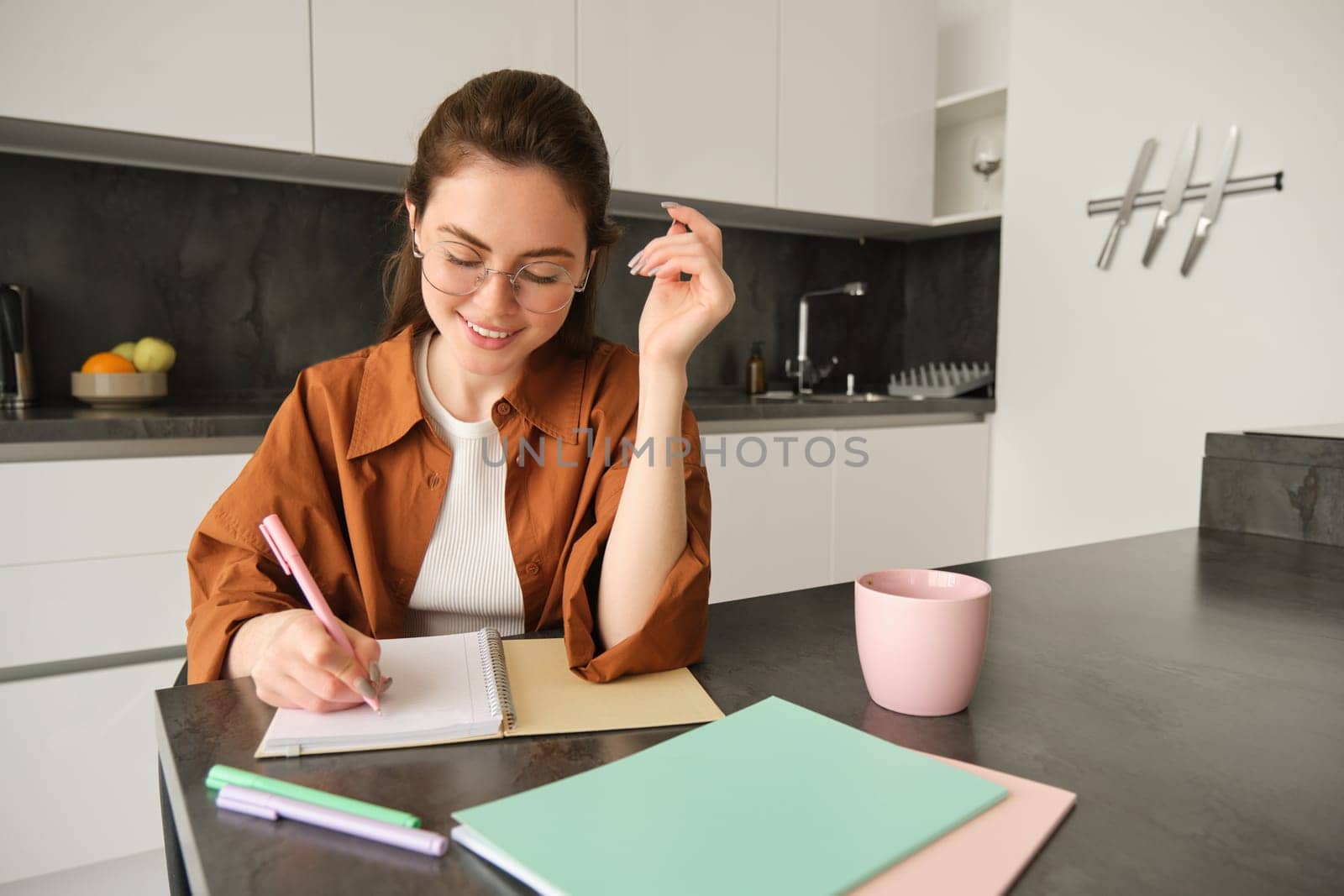 Portrait of young smiling woman, tutor preparing notes for lesson at home, writing in notebook and looking happy, student studying in kitchen.