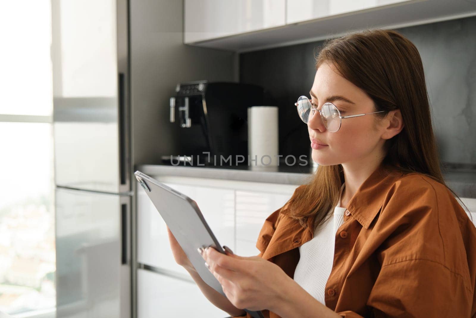 Side view of young beautiful woman spending time at home, reading digital tablet in glasses, sitting in kitchen.