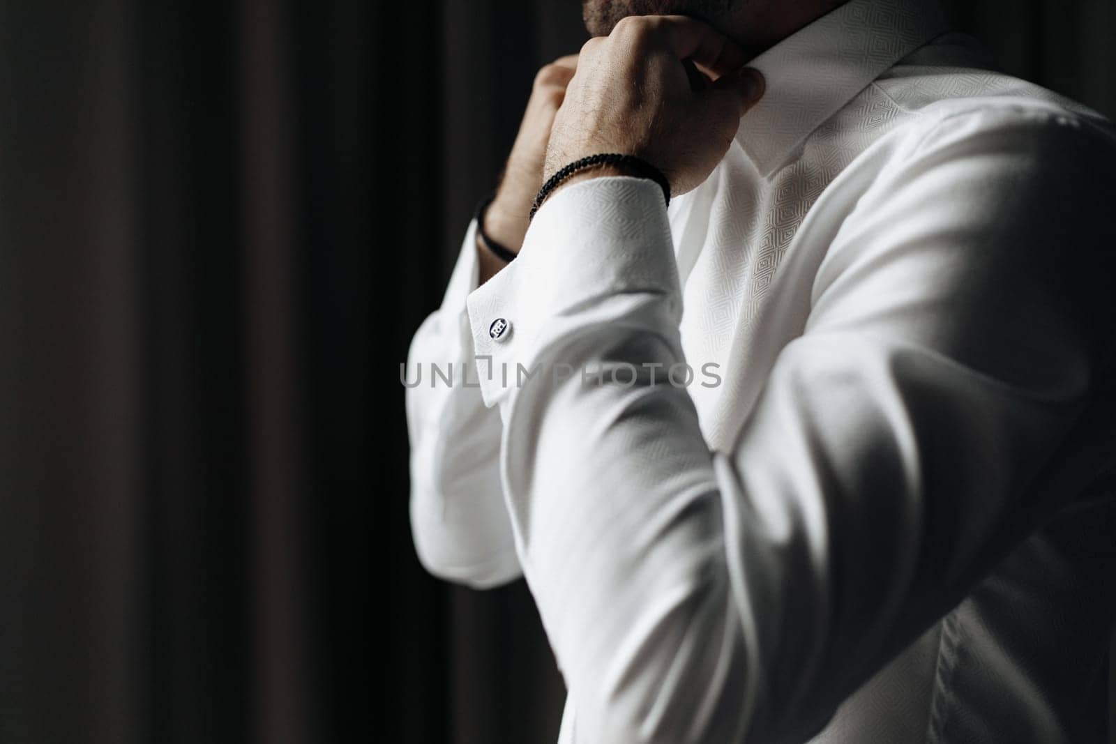 Close up cropped picture of caucasian male adjusting collar of a white shirt. wearing black bracelets. Groom preparing for wedding.Black curtain background copy-space.
