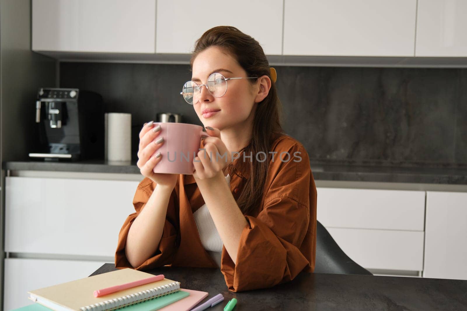 Portrait of young self-employed woman, working from home, taking break for coffee, sitting with documents in kitchen, looking away.