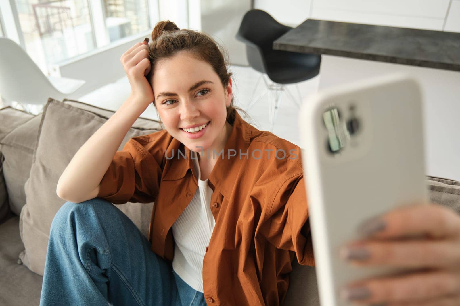 Carefree modern woman, smiling and taking selfies on smartphone app, posing for photo on her couch, sitting on sofa with mobile phone.