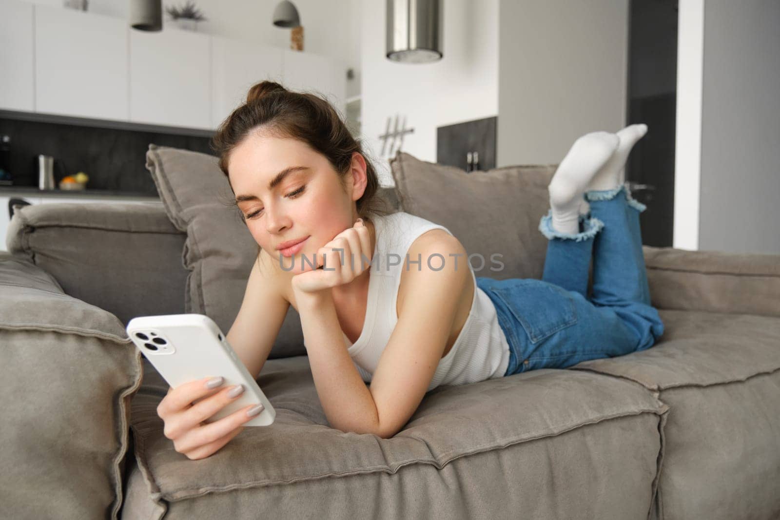 Portrait of smiling young woman lying on sofa at home, looking at her mobile phone, reading message on social media, using smartphone on couch.