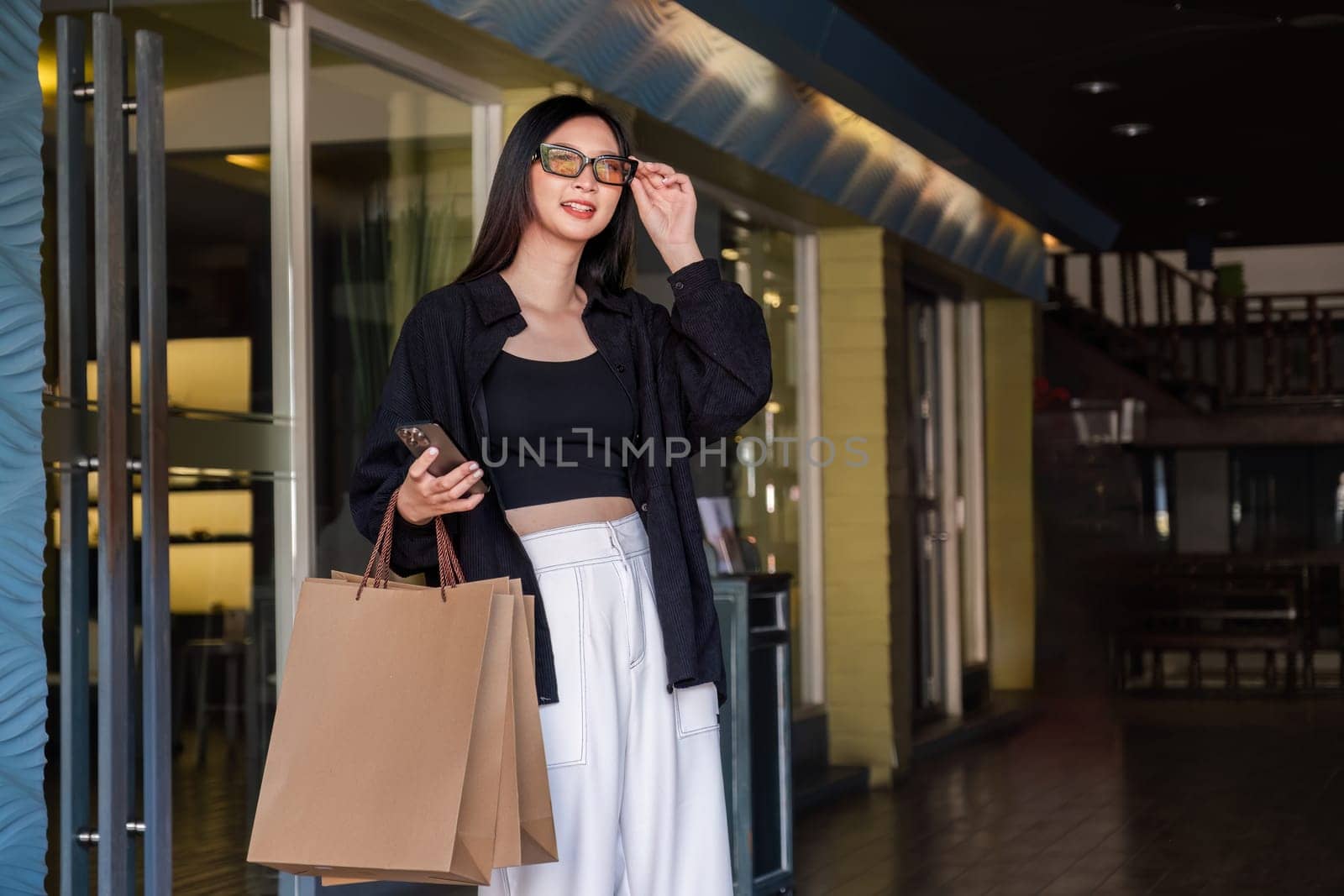 Asian woman using smartphone and looking away while enjoying a day shopping. Black Friday sale and discount. Buying clothes presents for holidays by wichayada