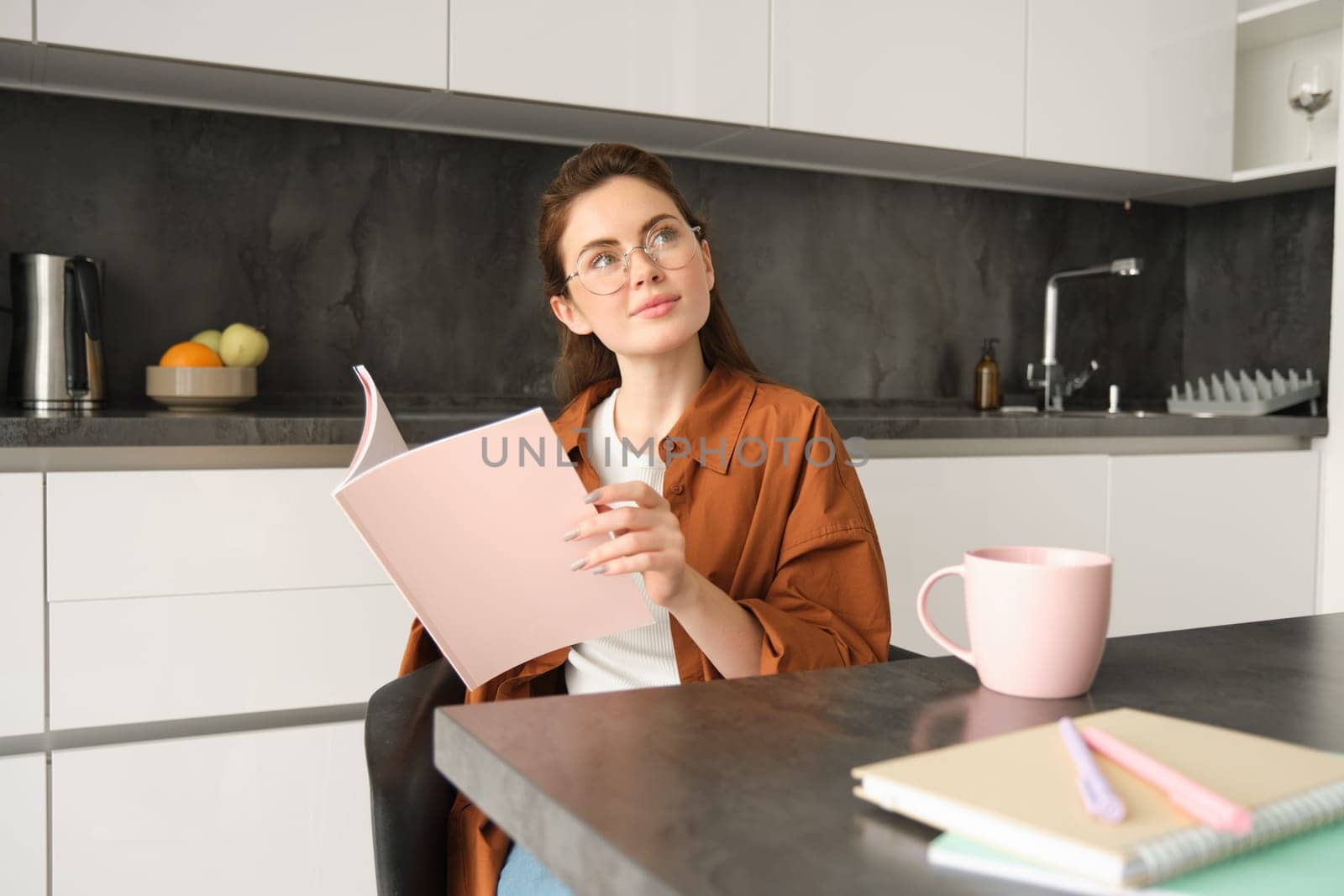 Portrait of young woman working from home, self-employed entrepreneur reading her documents, sitting at home in kitchen, wearing glasses.