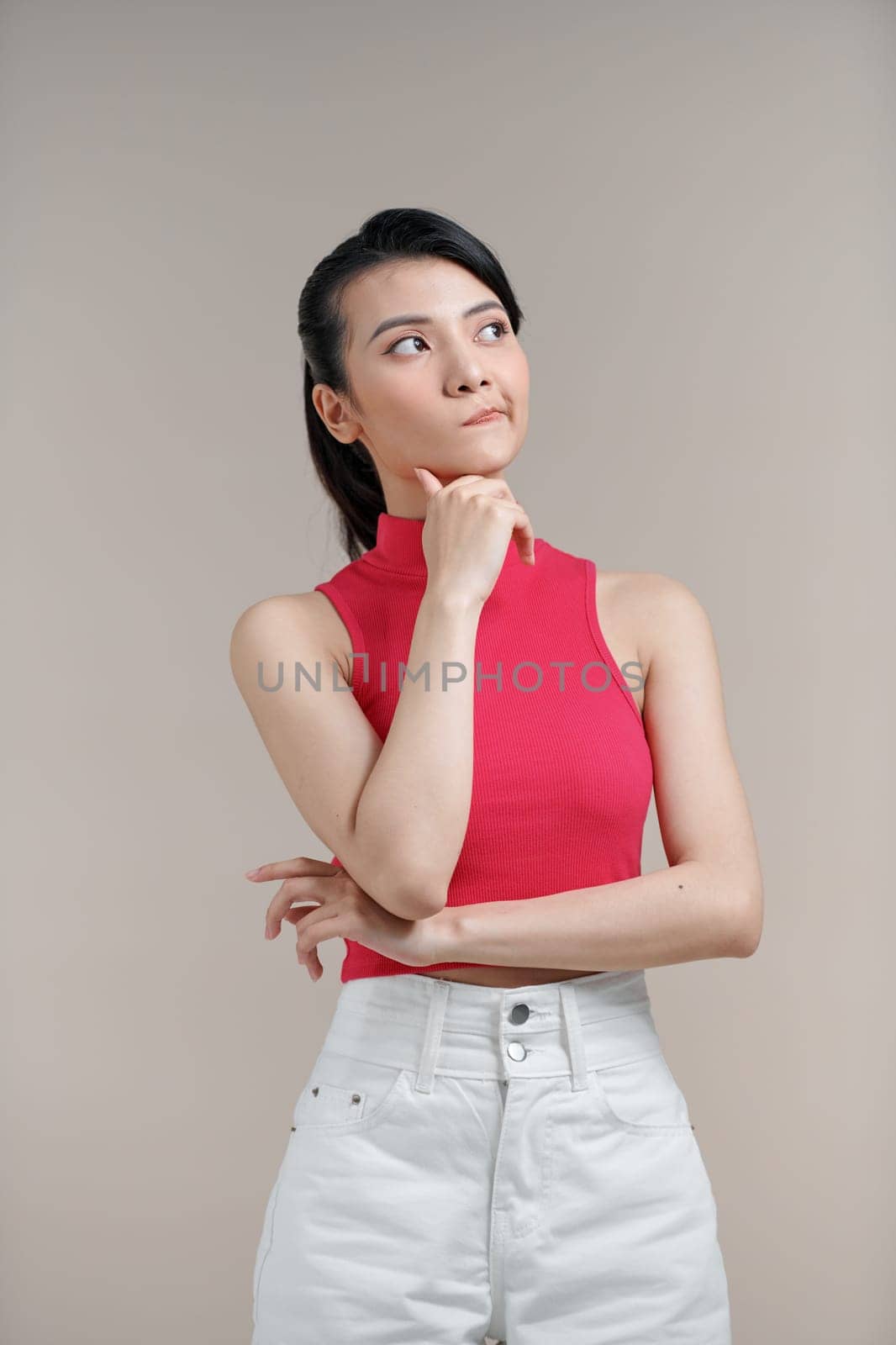 Asian thoughtful woman keeps hand on chin looks pensively against beige background