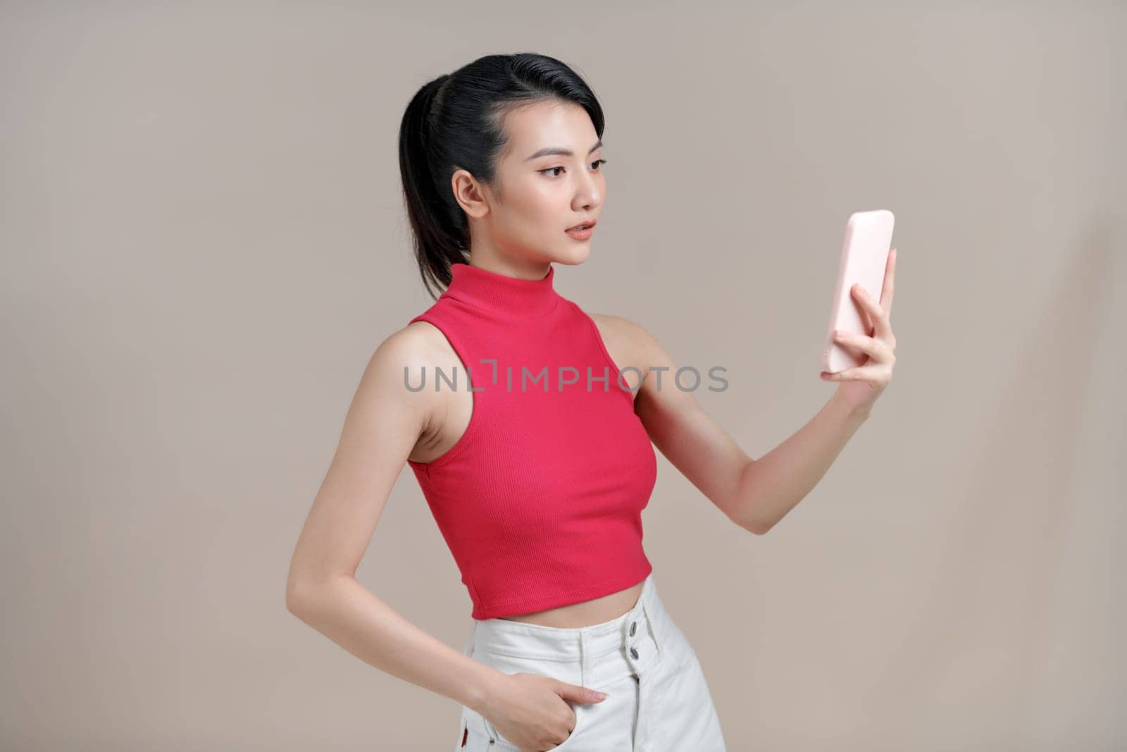 Beautiful young asian woman holding smartphone standing on isolated beige background.