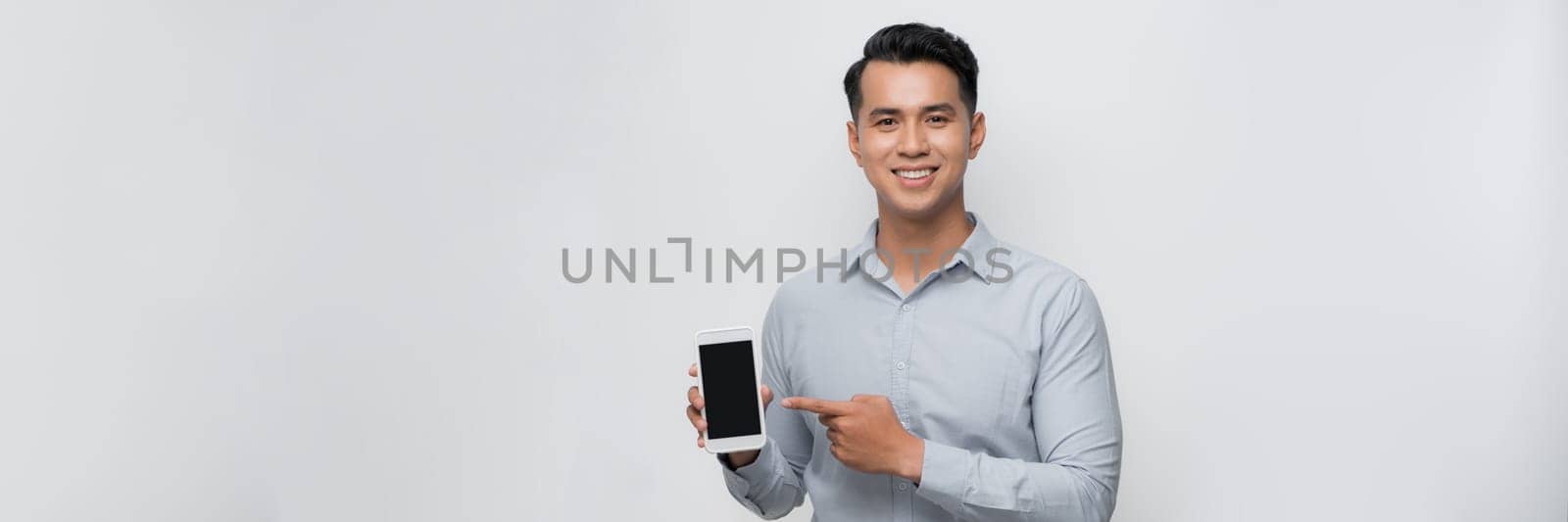 Smiling young man holds a modern smartphone in his hand and shows his finger on a blank screen by makidotvn