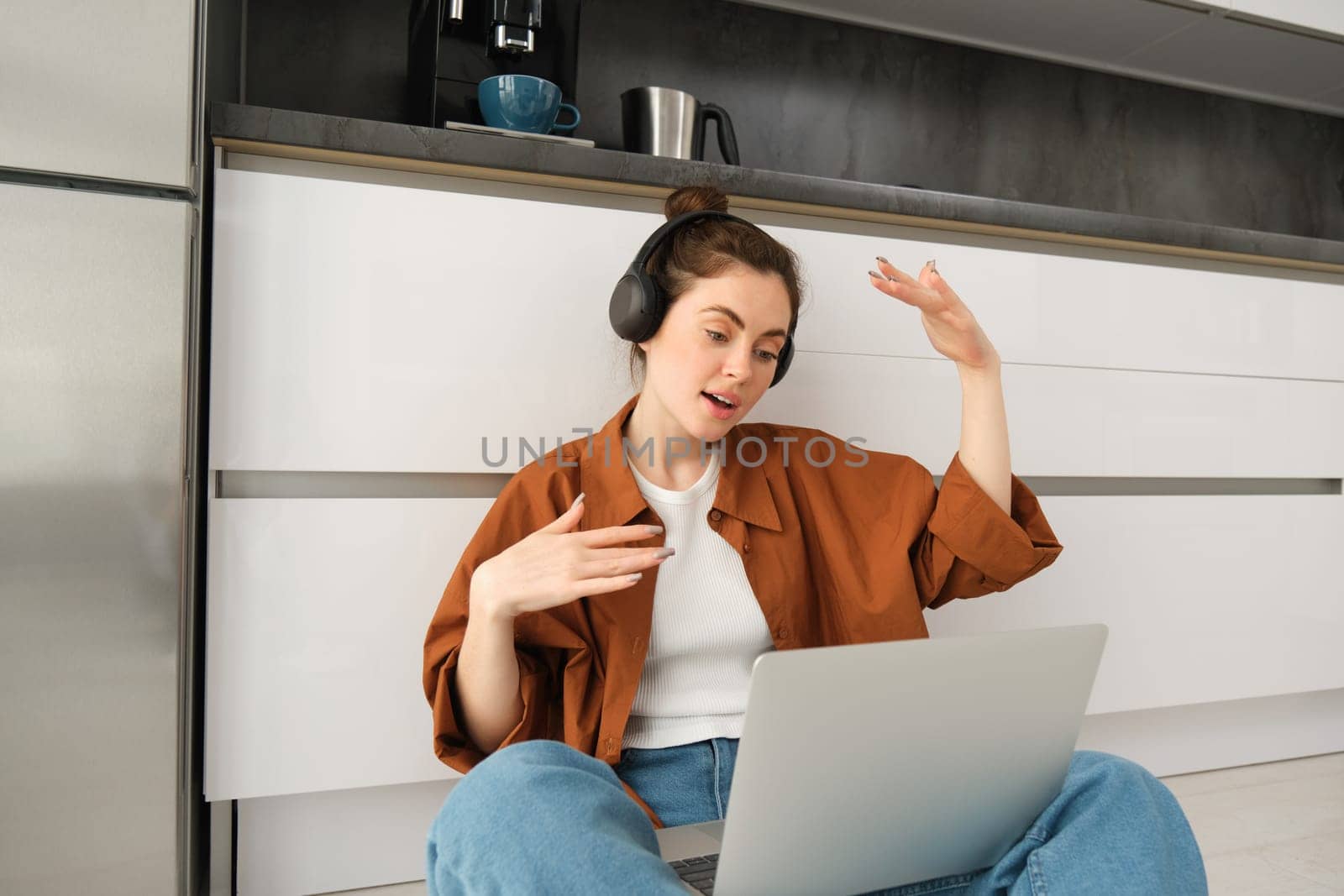 Portrait of woman listens to music in headphones, while working on laptop, sits at home on kitchen floor, making dance movements.