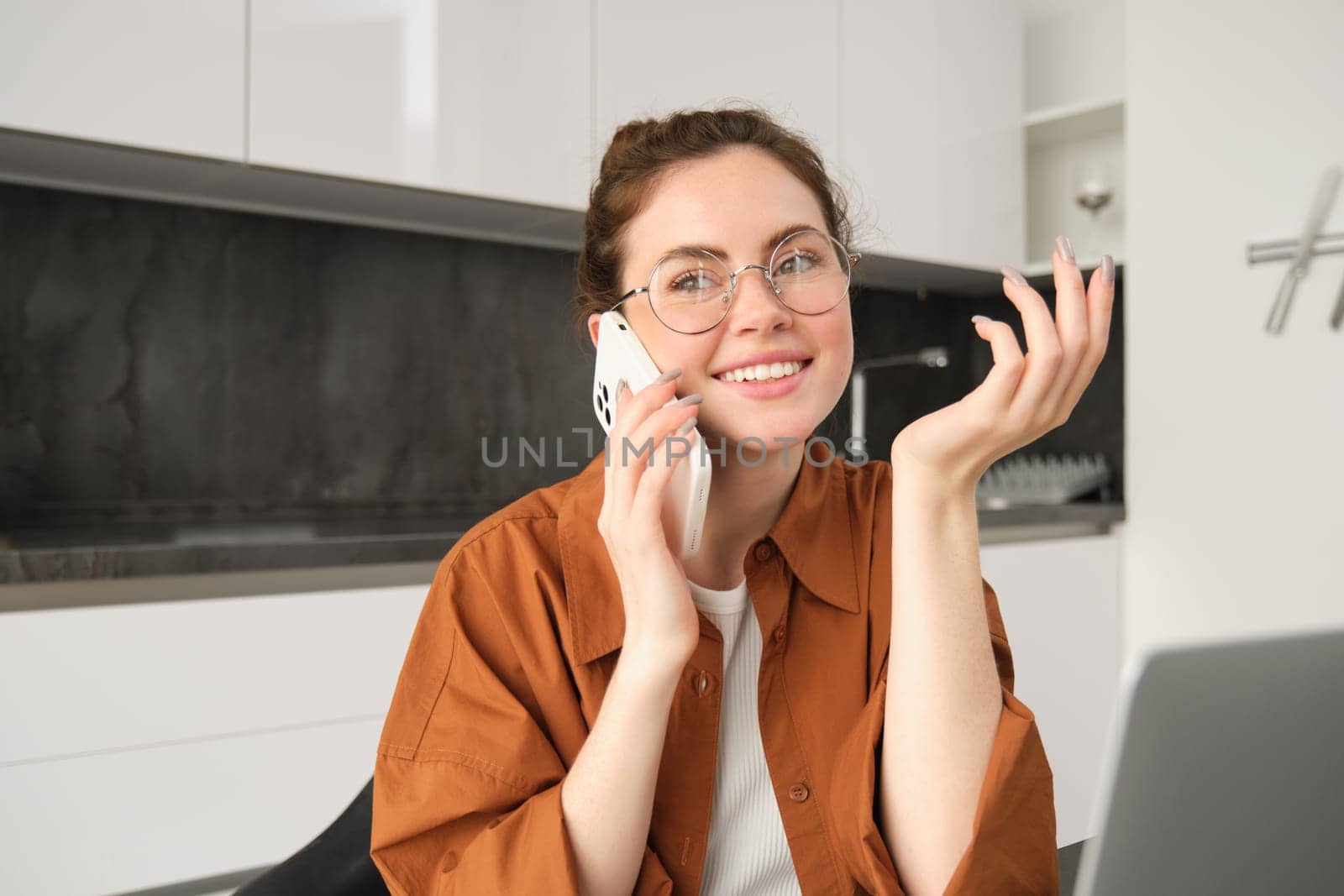 Portrait of young woman, business owner working from home, student making a phone call, sitting in kitchen with laptop, talking to someone.