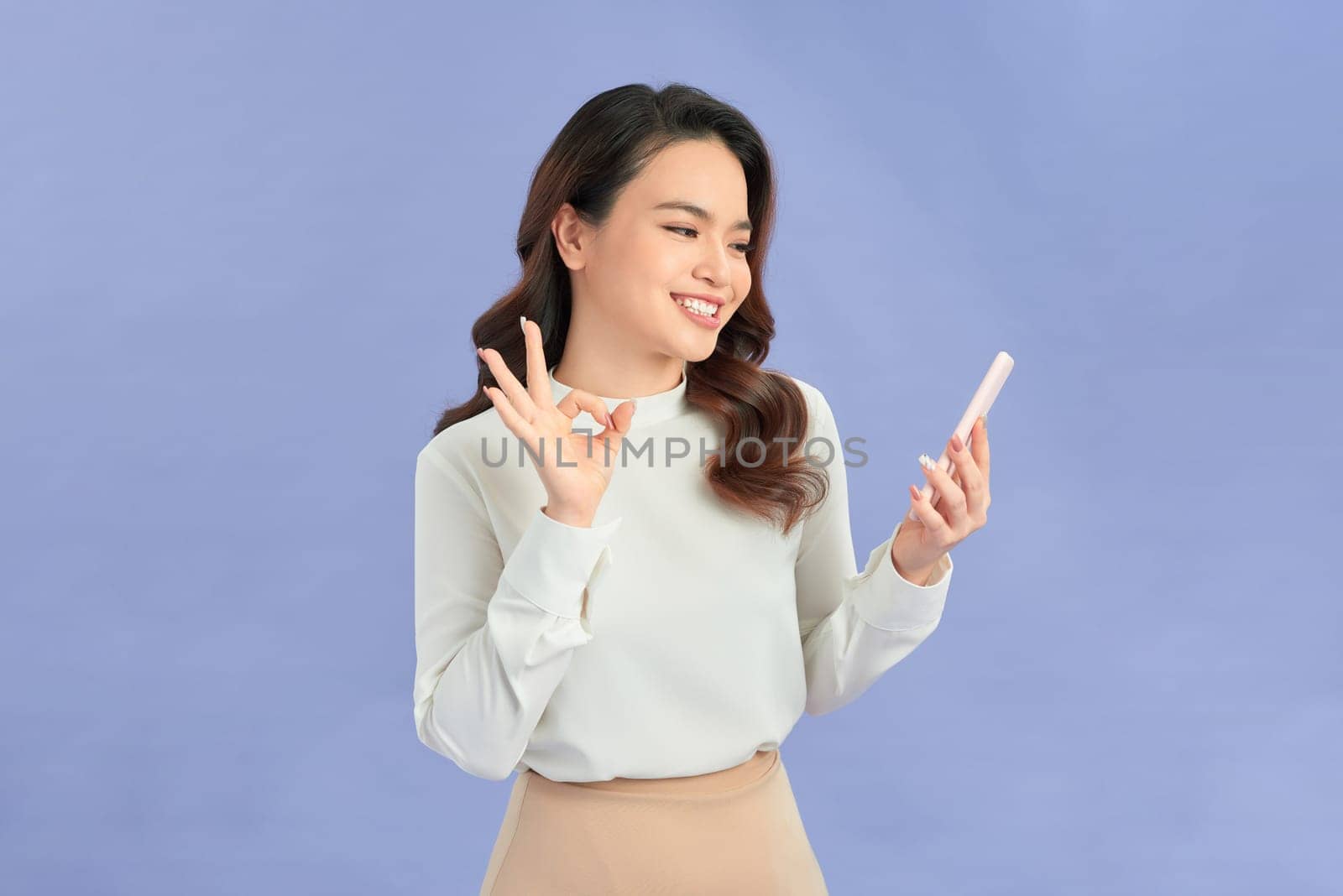 Cheerful Asian woman holding smartphone and shows ok sign on light violet background. by makidotvn
