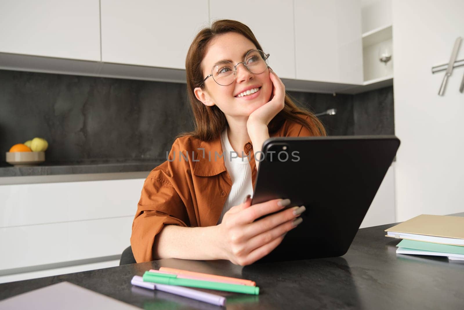 Portrait of young student, woman studying at home, working remotely, setup workplace in her kitchen, sitting on chair with digital tablet, reading in glasses.